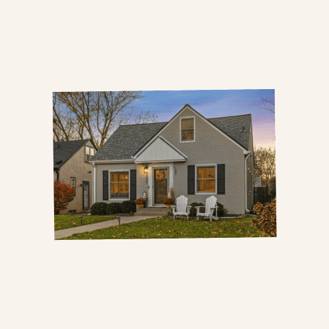 Charming two-story house with beige siding, a sloped roof, and white trim, featuring two front windows and a central front door. The house is surrounded by a well-kept lawn and pathway, flanked by two white Adirondack chairs and shrubs. The background shows trees and a sunset sky.