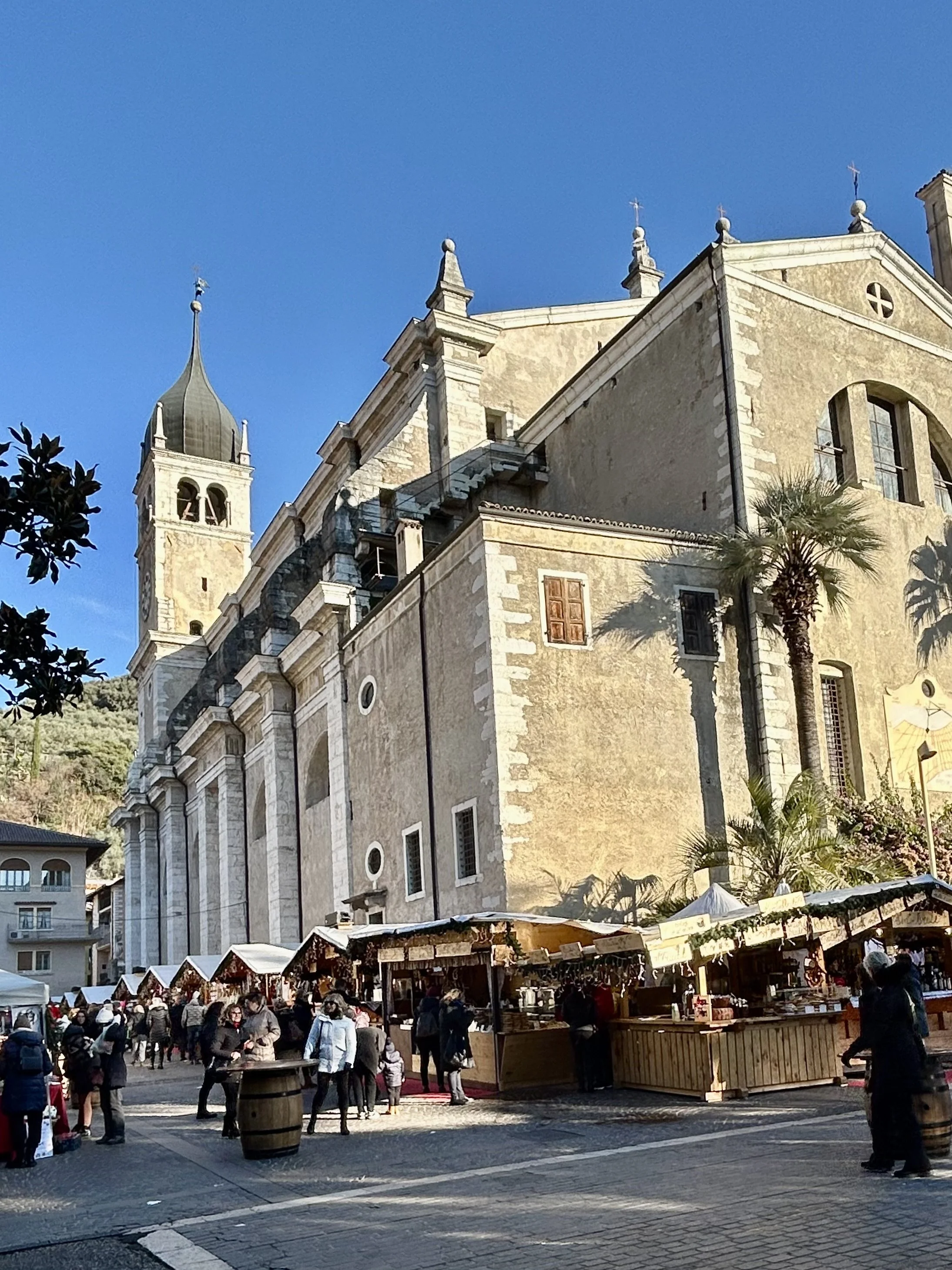 A Christmas market set up in front of a historic stone church near Lake Garda, with wooden stalls, palm trees, and visitors walking among the booths under a bright blue sky.