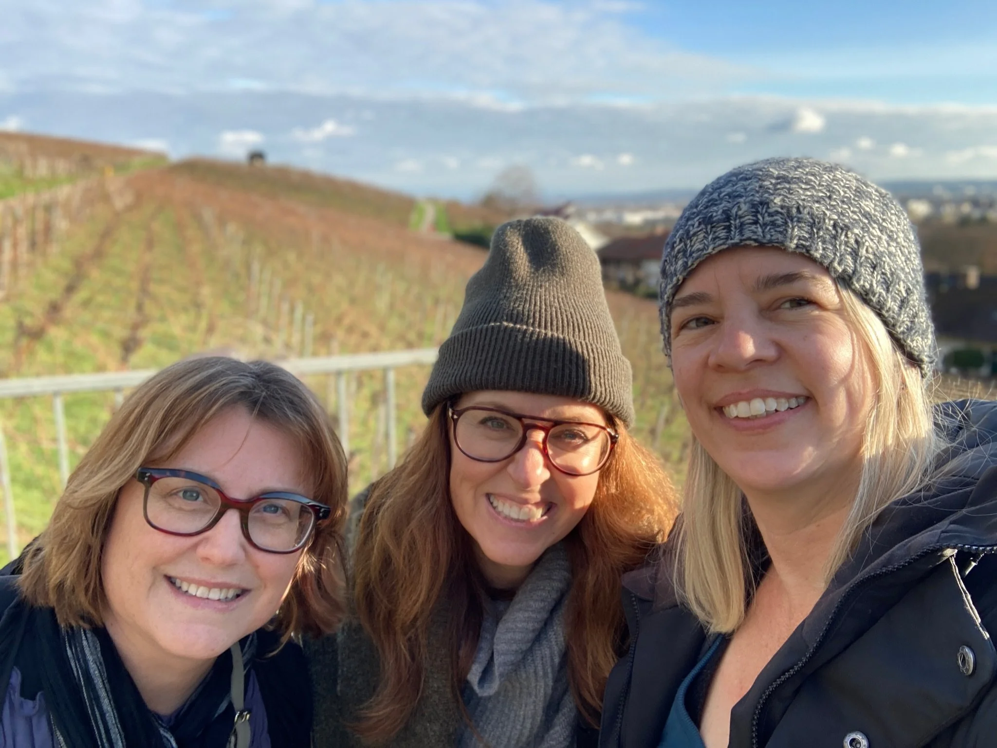 Three women smiling in front of winter vineyards in the Alsace wine region under a bright, partly cloudy sky.