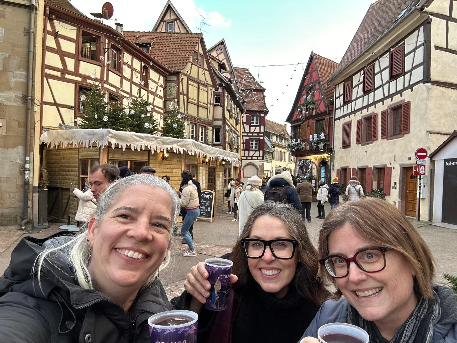 Three women holding mulled wine cups in front of colorful half-timbered buildings at the Colmar Christmas market, with festive decorations and people exploring the old town.