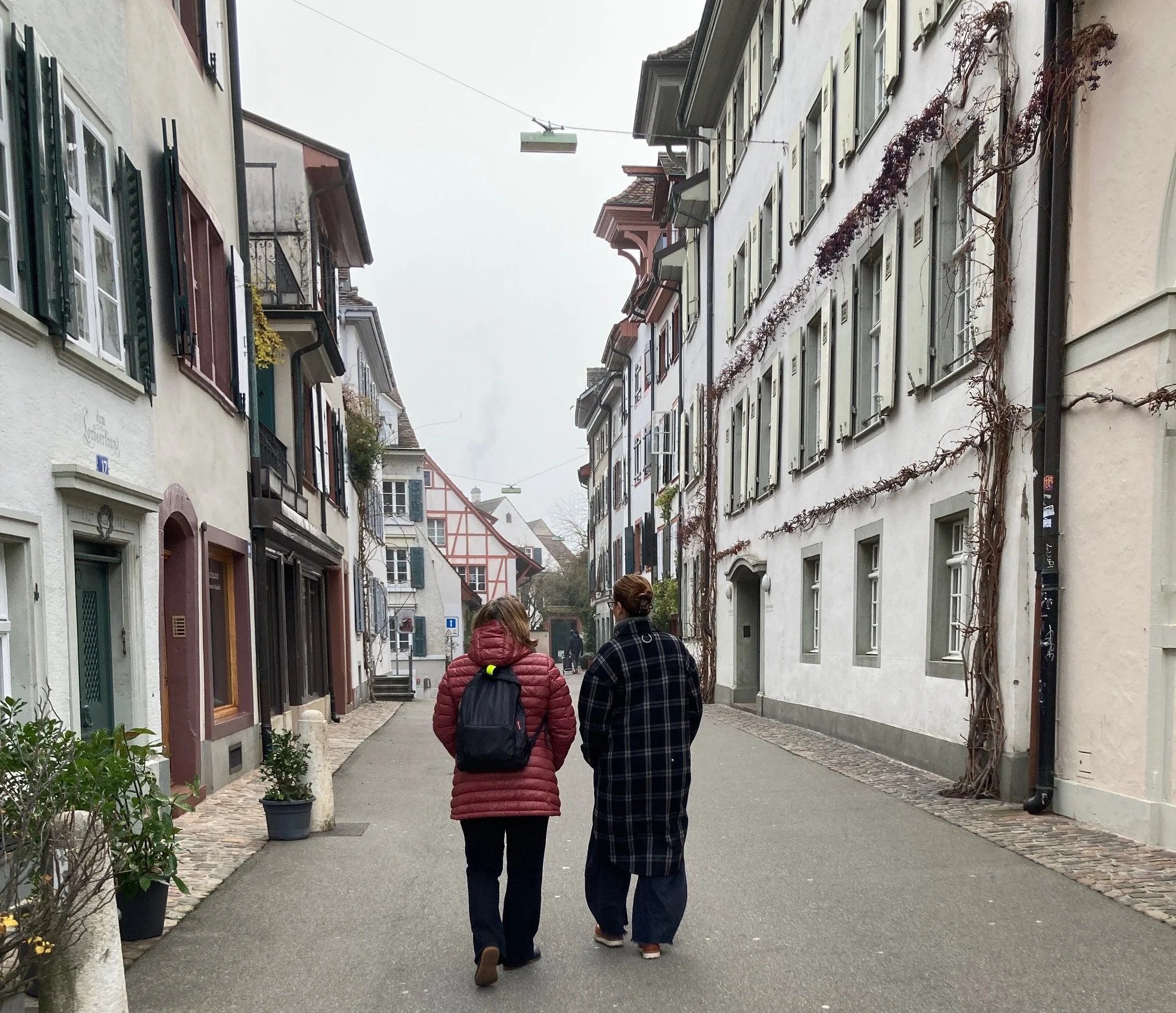 Two women walking down a quiet, foggy street in Basel’s old town, lined with pastel buildings, shutters, cobblestones, and winter vines on the façades.