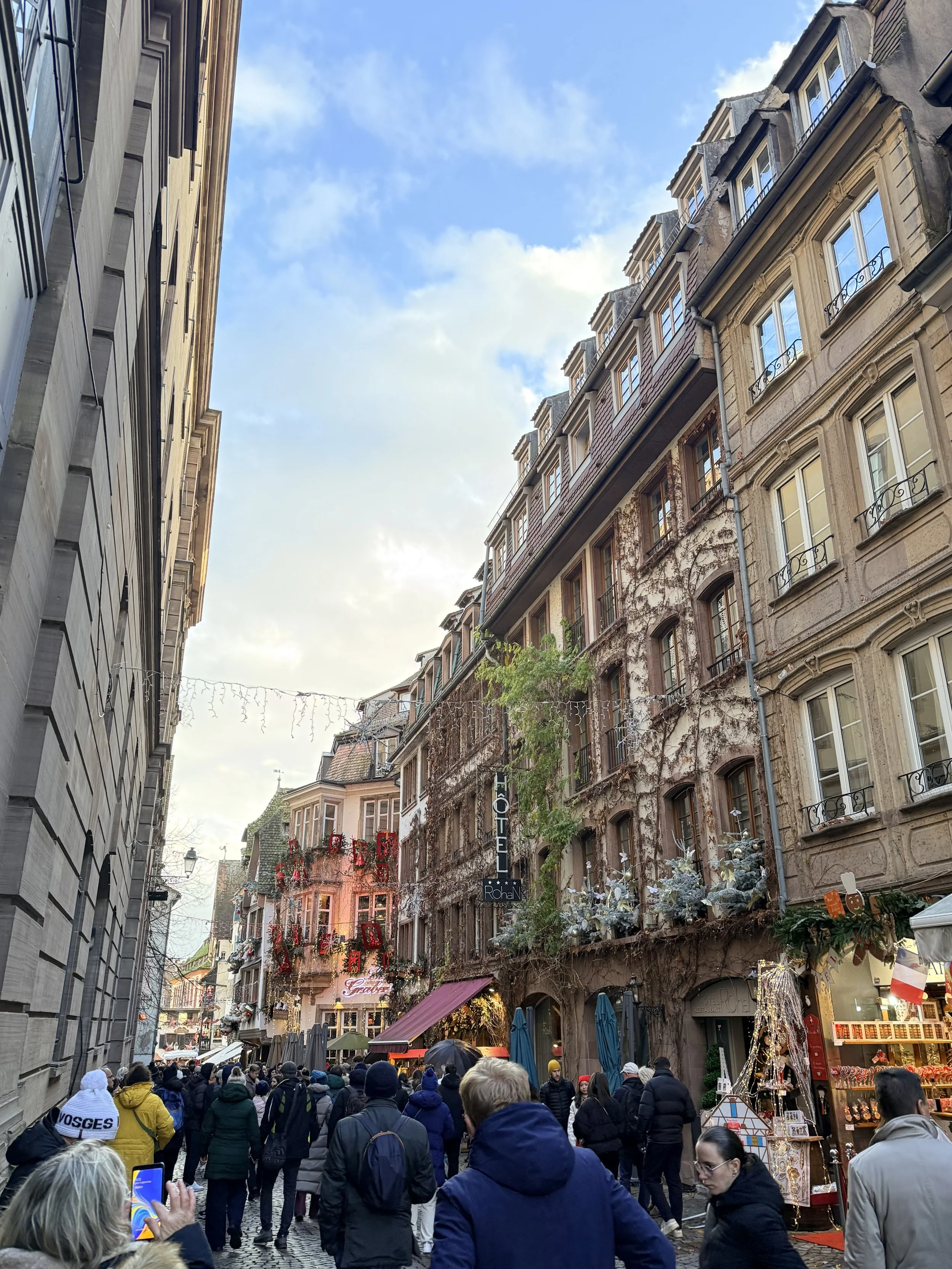 Crowded pedestrian street in Strasbourg decorated with holiday lights, with historic buildings, shop displays, and tourists exploring the Christmas markets.