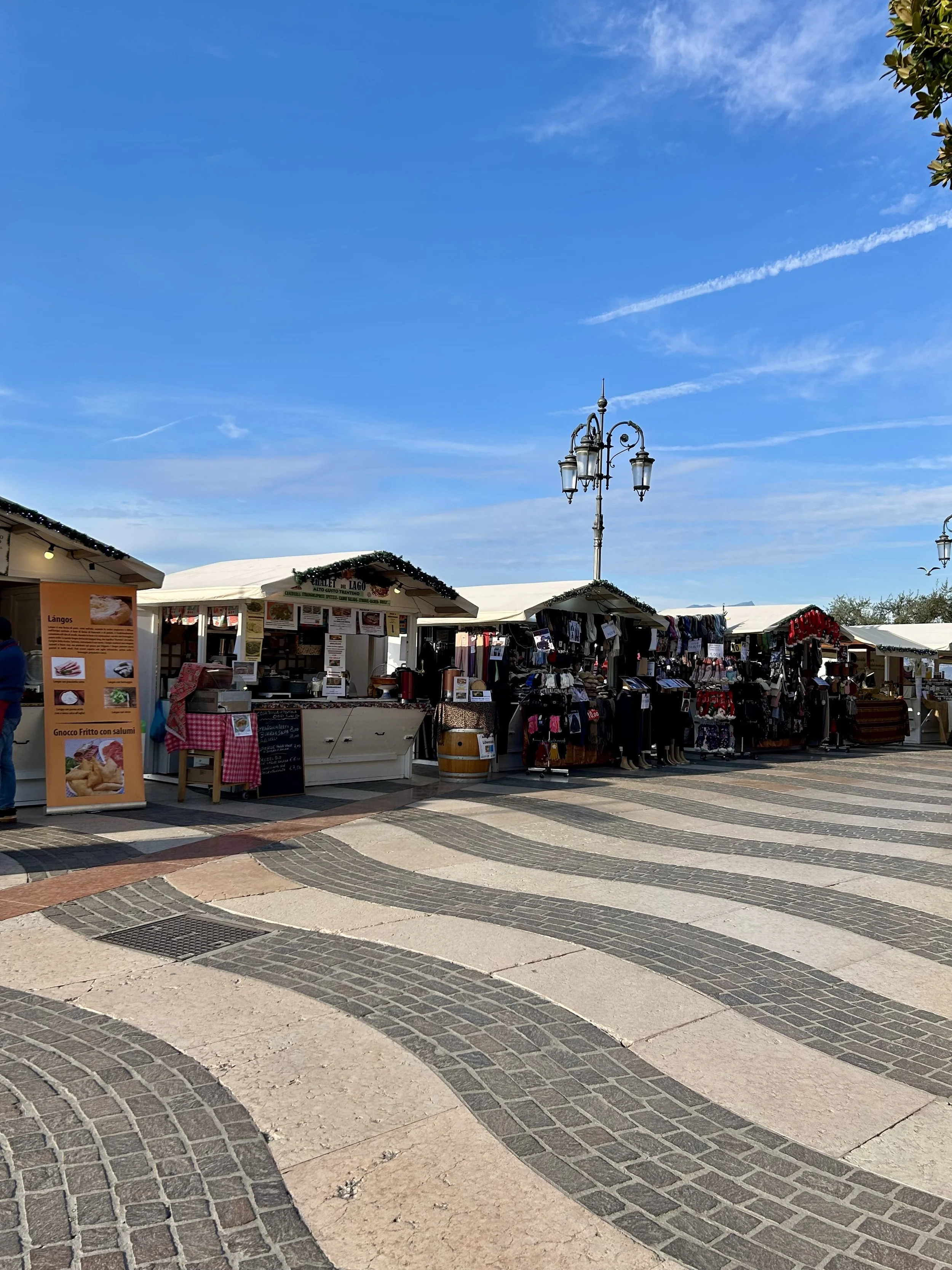 Christmas market stalls lined up along a lakeside promenade under a bright blue sky in Italy