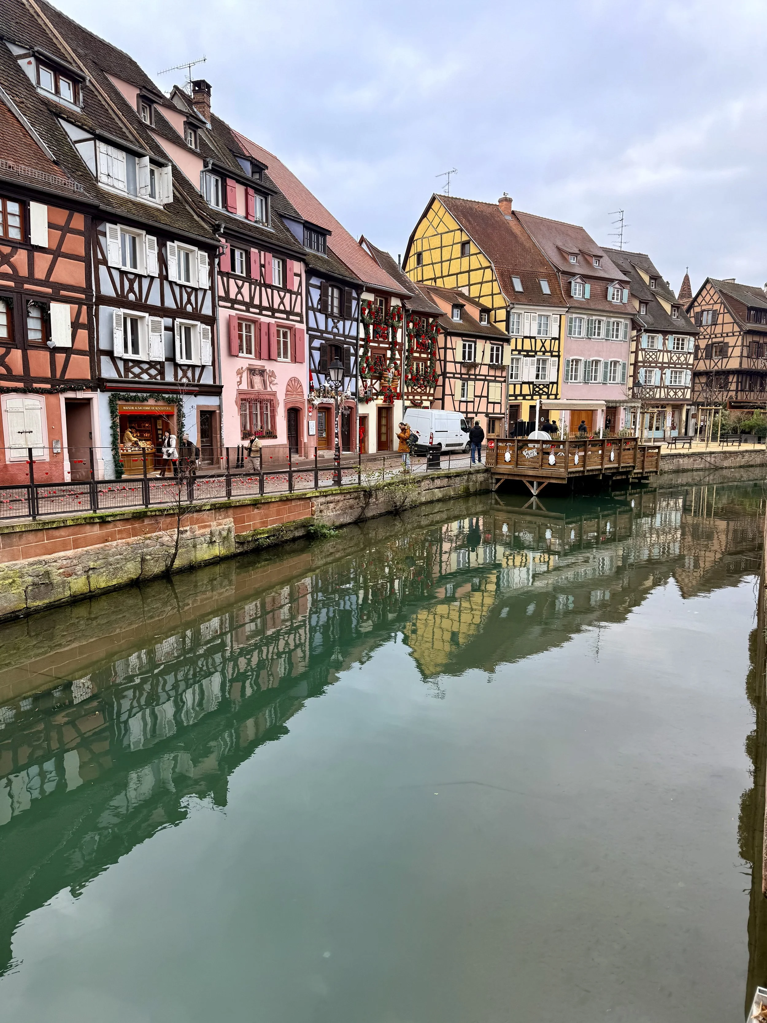 Colorful half-timbered houses reflecting in the calm canal in Colmar’s Little Venice district on a cloudy day.