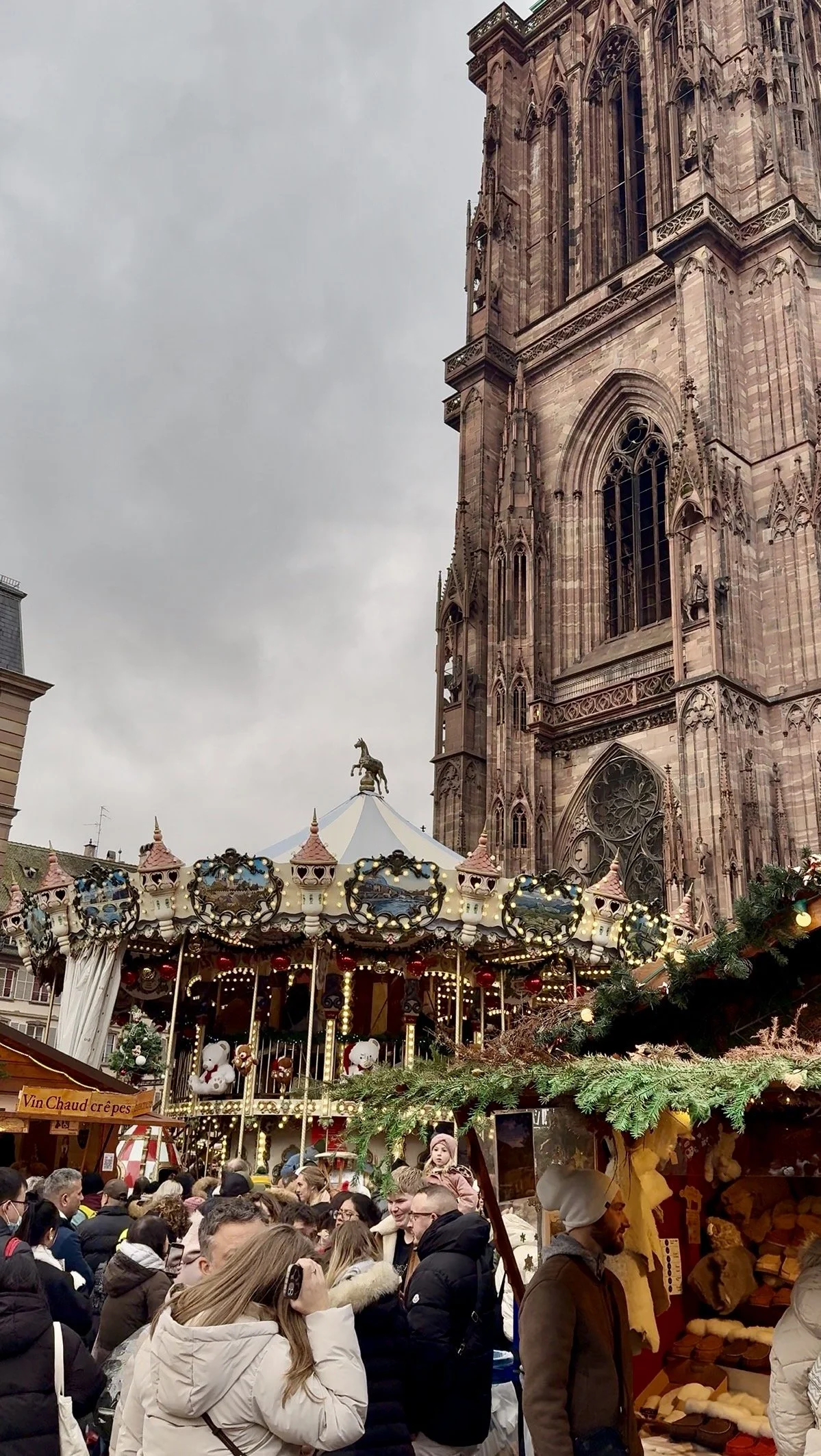Festive Christmas carousel in front of Strasbourg Cathedral, surrounded by holiday lights, evergreen garlands, and crowds of visitors at the market.