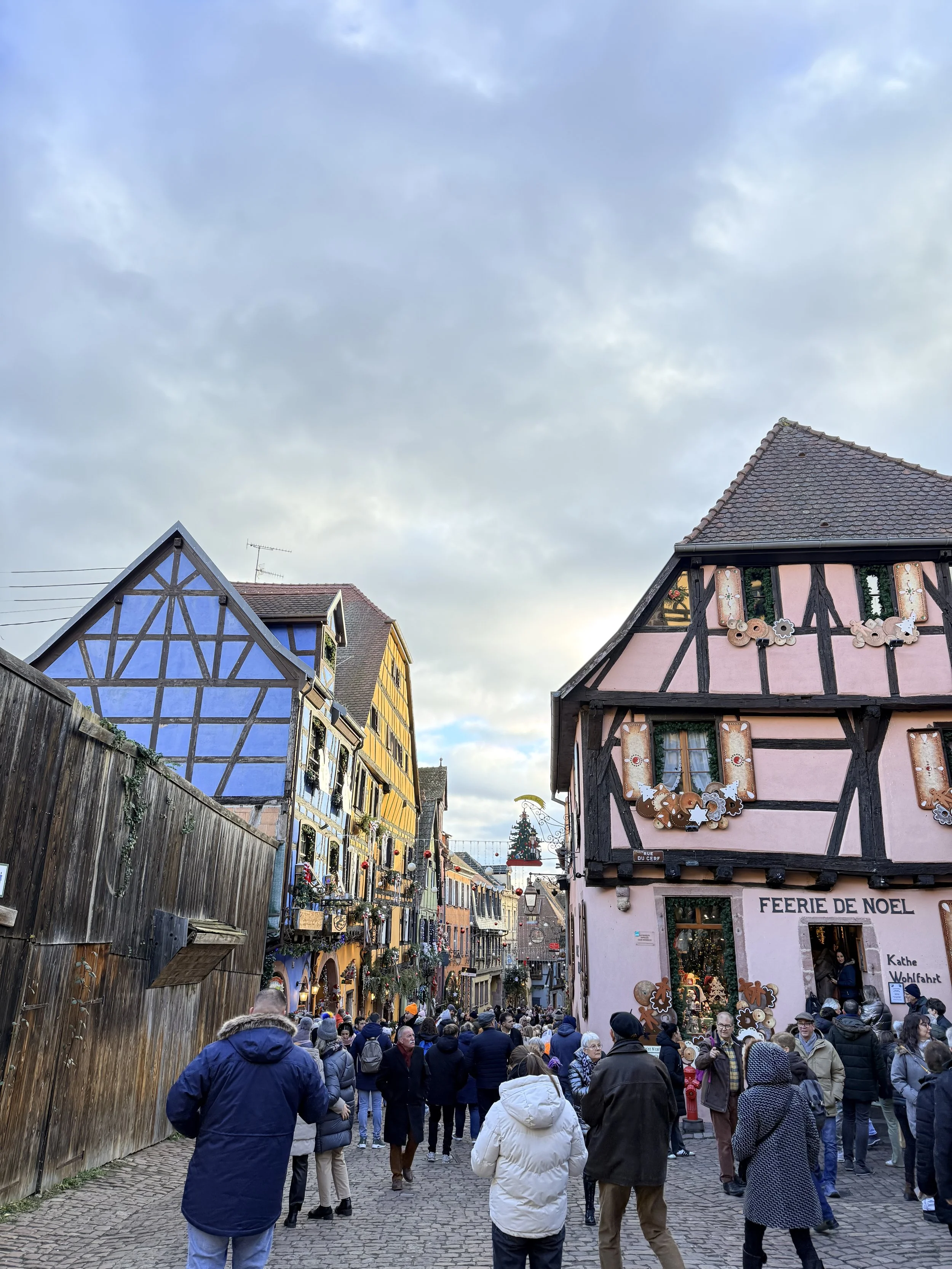 A picturesque cobblestone street in Riquewihr lined with colorful half-timbered homes covered in Christmas decorations, with crowds walking toward the market.