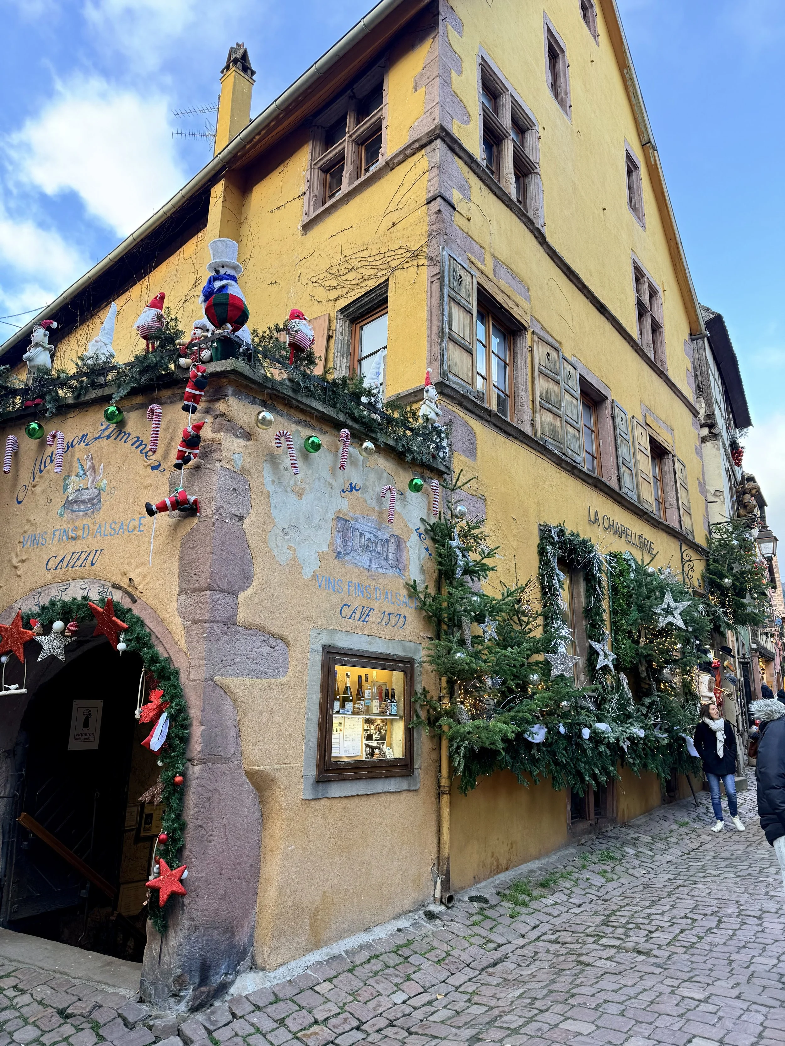 A brightly painted yellow medieval building in Riquewihr covered in whimsical Christmas decorations, garlands, and ornaments, with cobblestone streets below.
