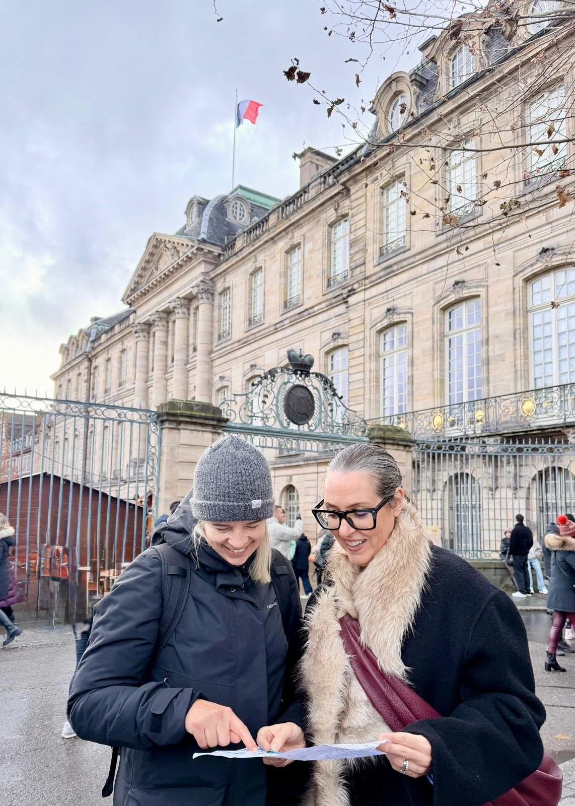 Two women looking at a map and laughing in front of the ornate Palais Rohan in Strasbourg, with winter coats, a French flag flying above the building, and Christmas market stalls nearby.