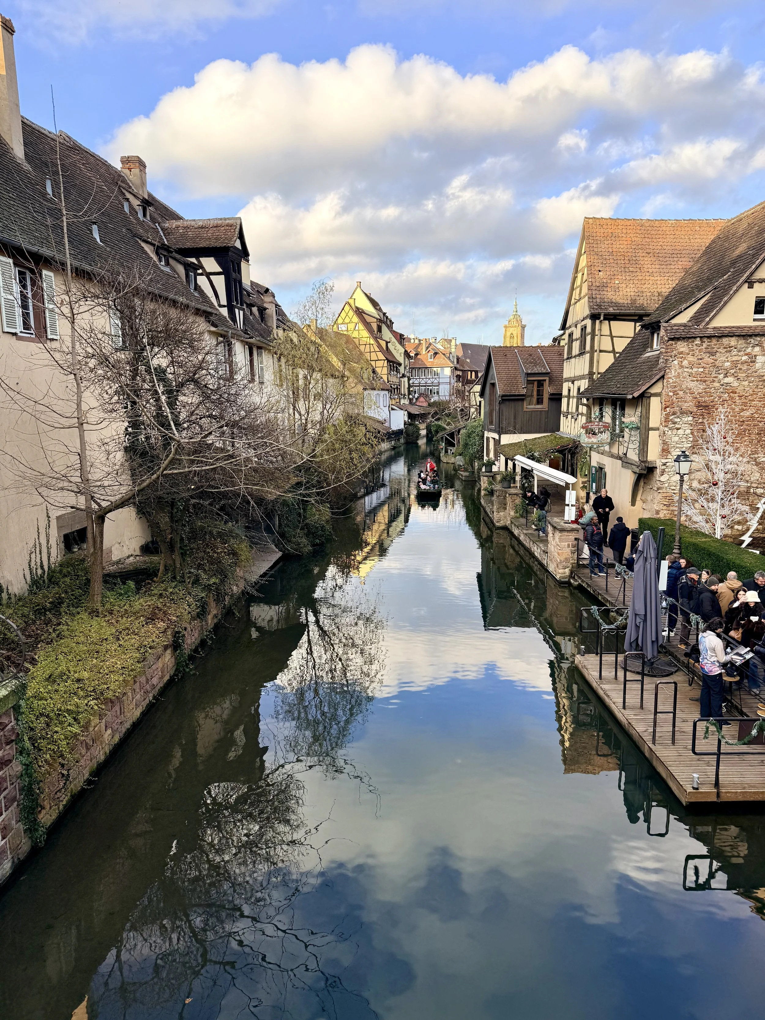 A peaceful canal in Colmar lined with historic half-timbered buildings, reflecting the sky and clouds on the water’s surface