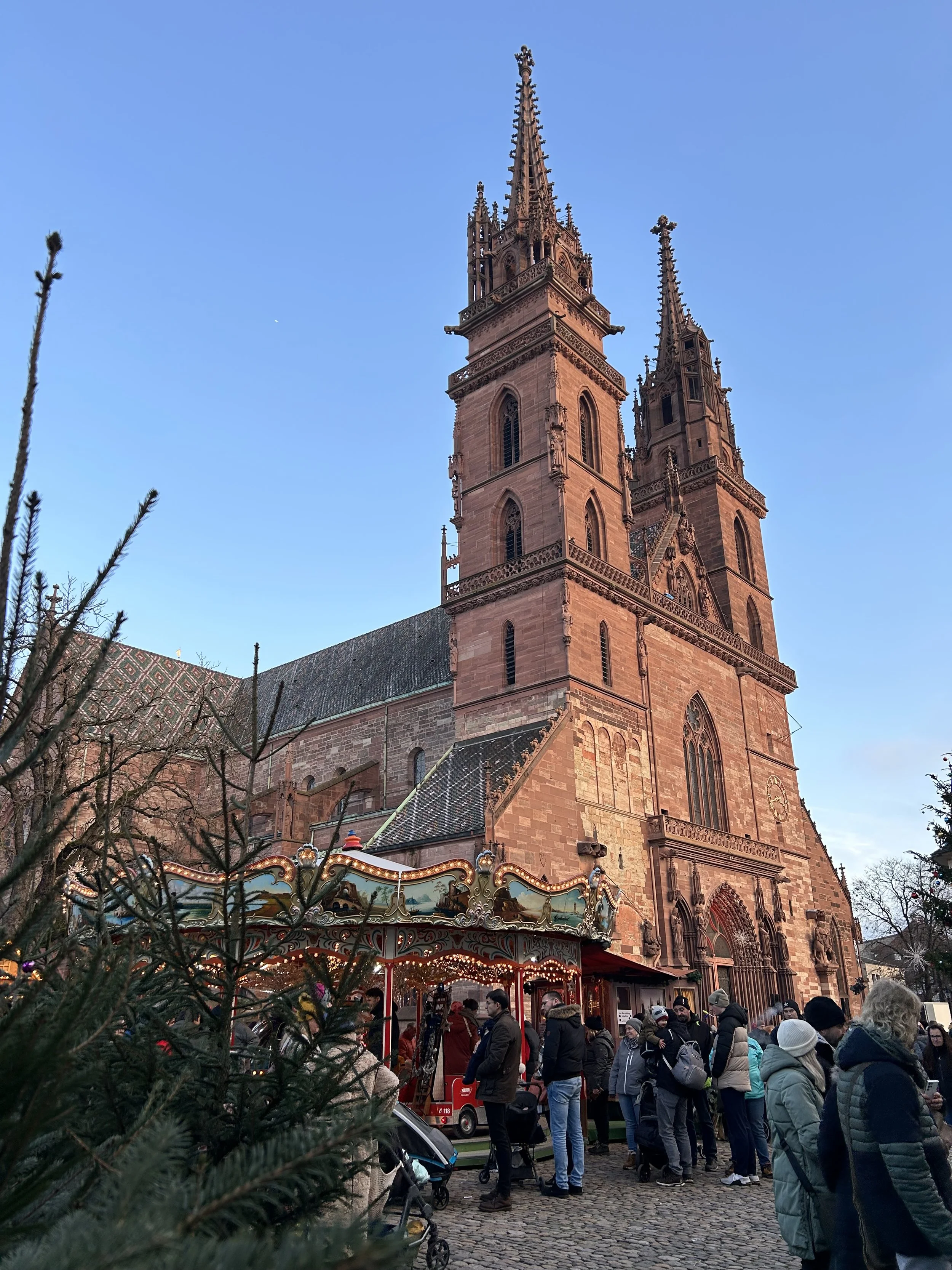 A Christmas carousel next to the red sandstone Basel Cathedral with people strolling through the market stalls