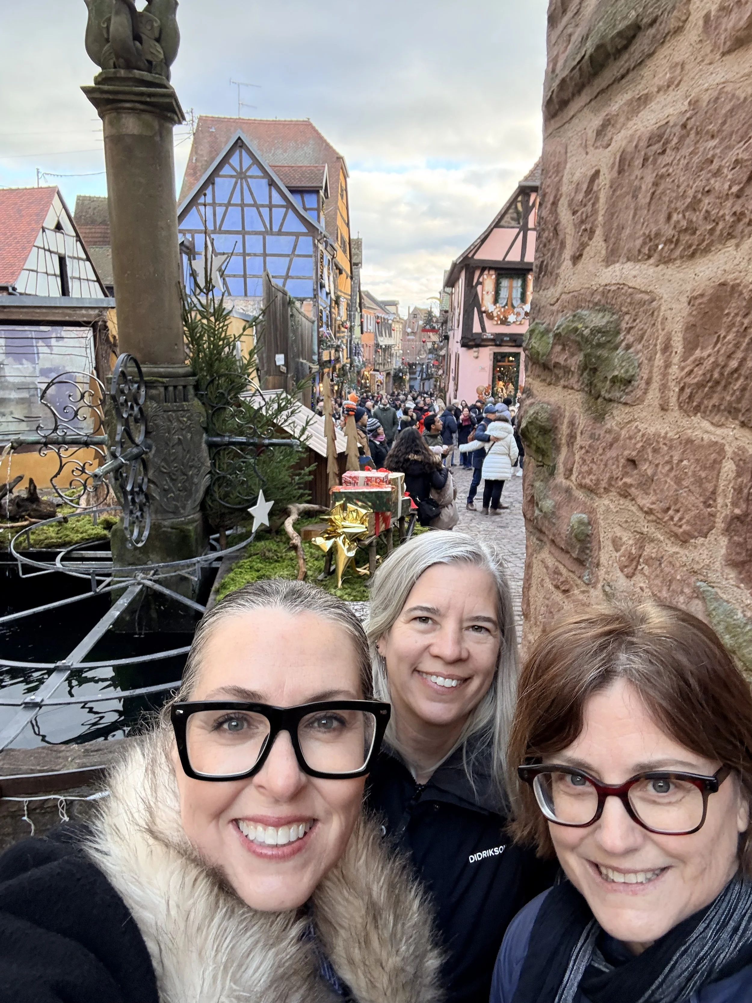 Selfie of three women smiling in front of Riquewihr’s half-timbered houses and Christmas market crowds on a festive winter day.