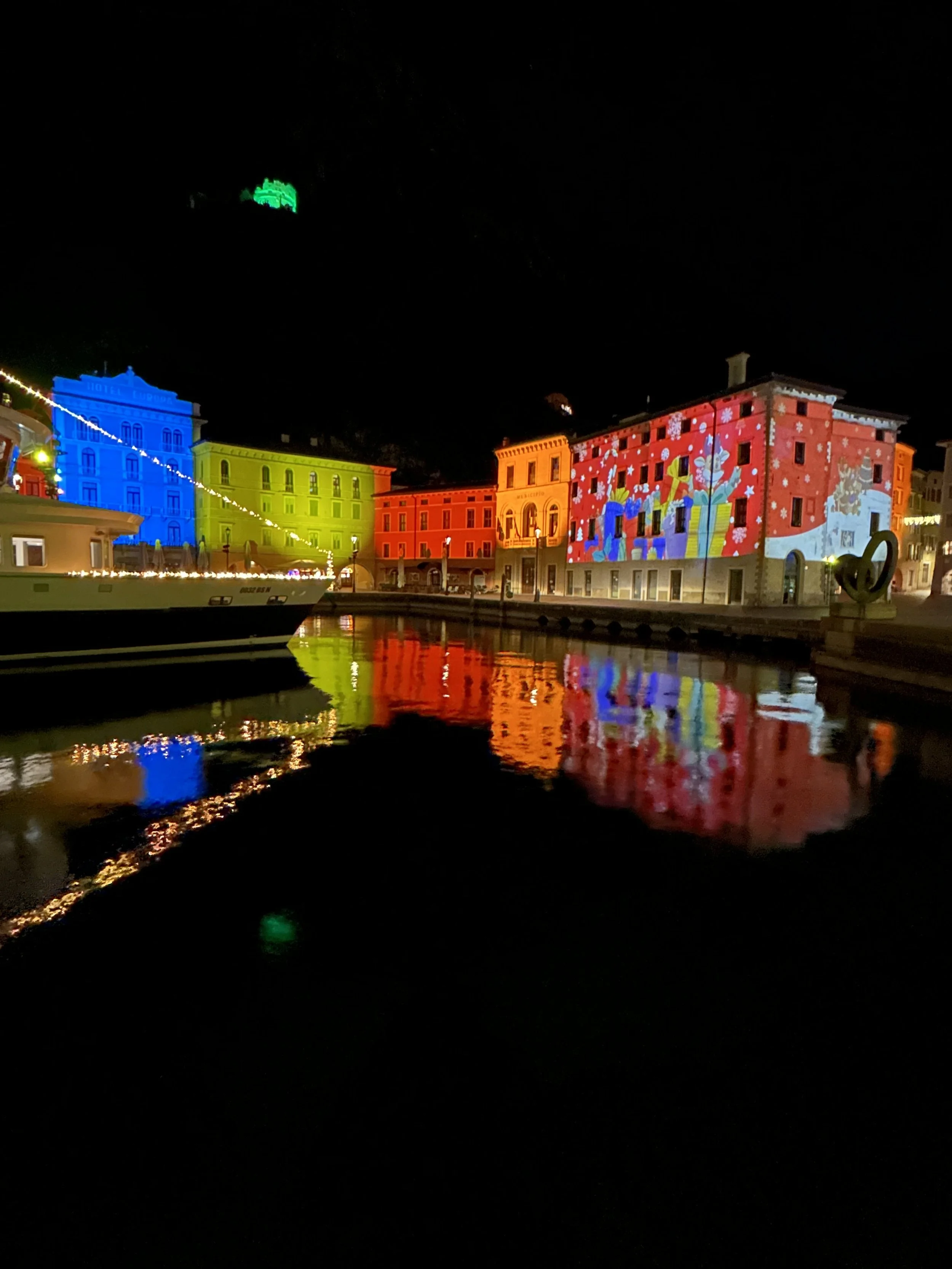 Vibrant Christmas light projections illuminating the waterfront buildings in Riva del Garda, reflecting across the dark lake at night