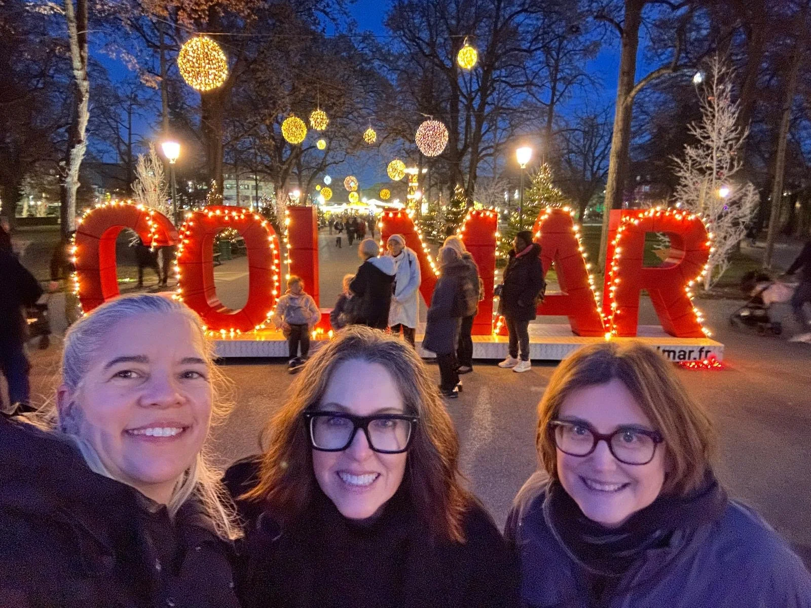 Three women smiling in front of a large illuminated “COLMAR” sign at a Christmas market at dusk, with glowing ornaments hanging from the trees and people walking in the background