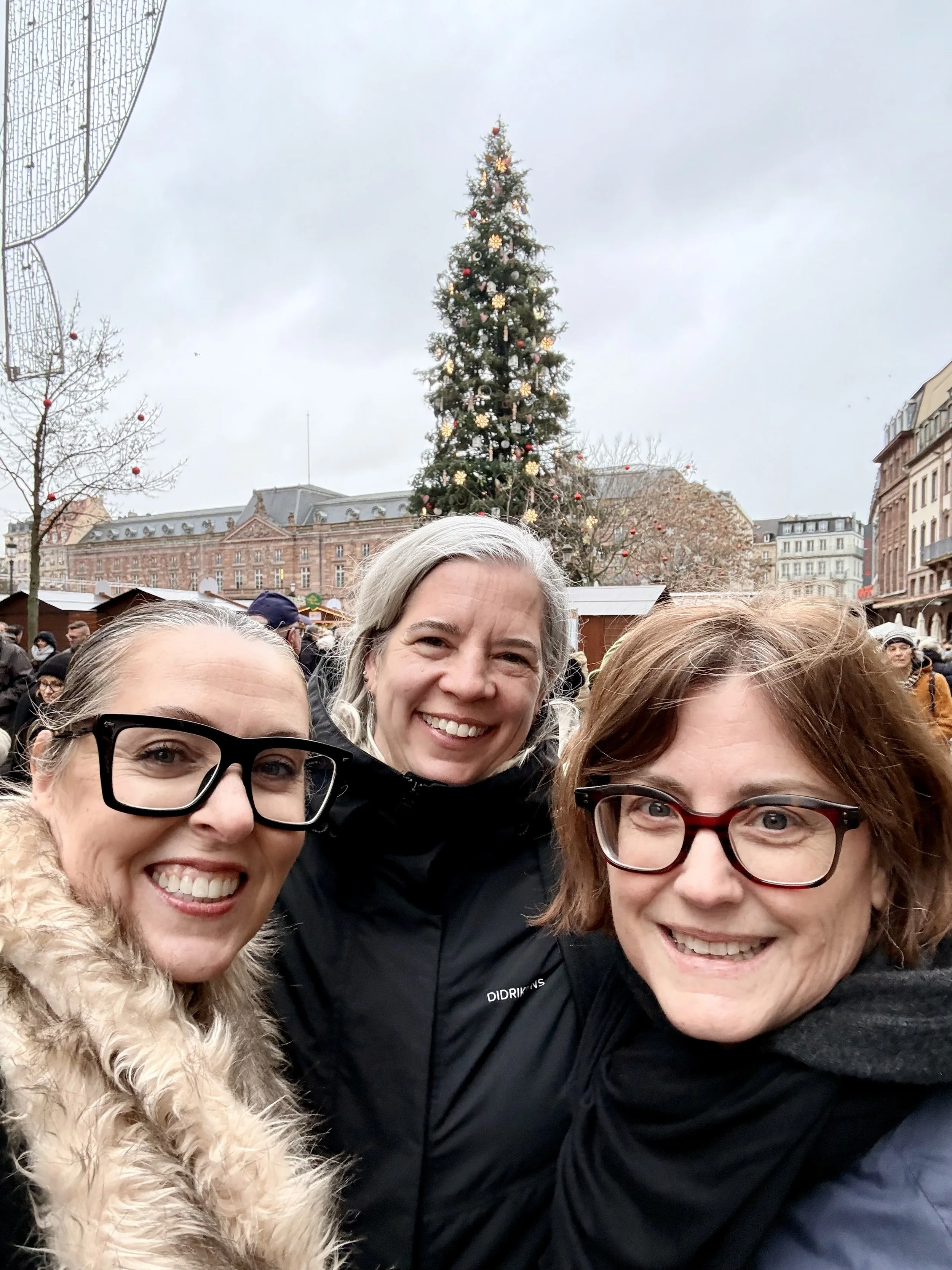 Three women smiling in front of Strasbourg’s large decorated Christmas tree in the central square, surrounded by crowds and historic buildings