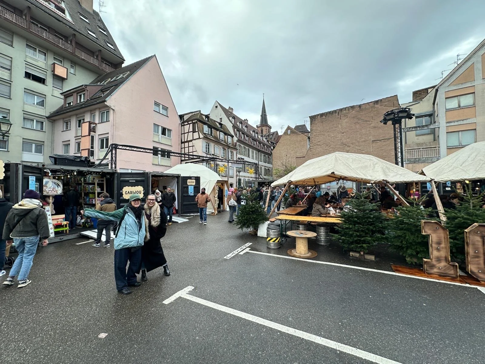 Outdoor Christmas market in Strasbourg with wooden stalls and covered seating tents, surrounded by pastel buildings and crowds browsing crafts and food.