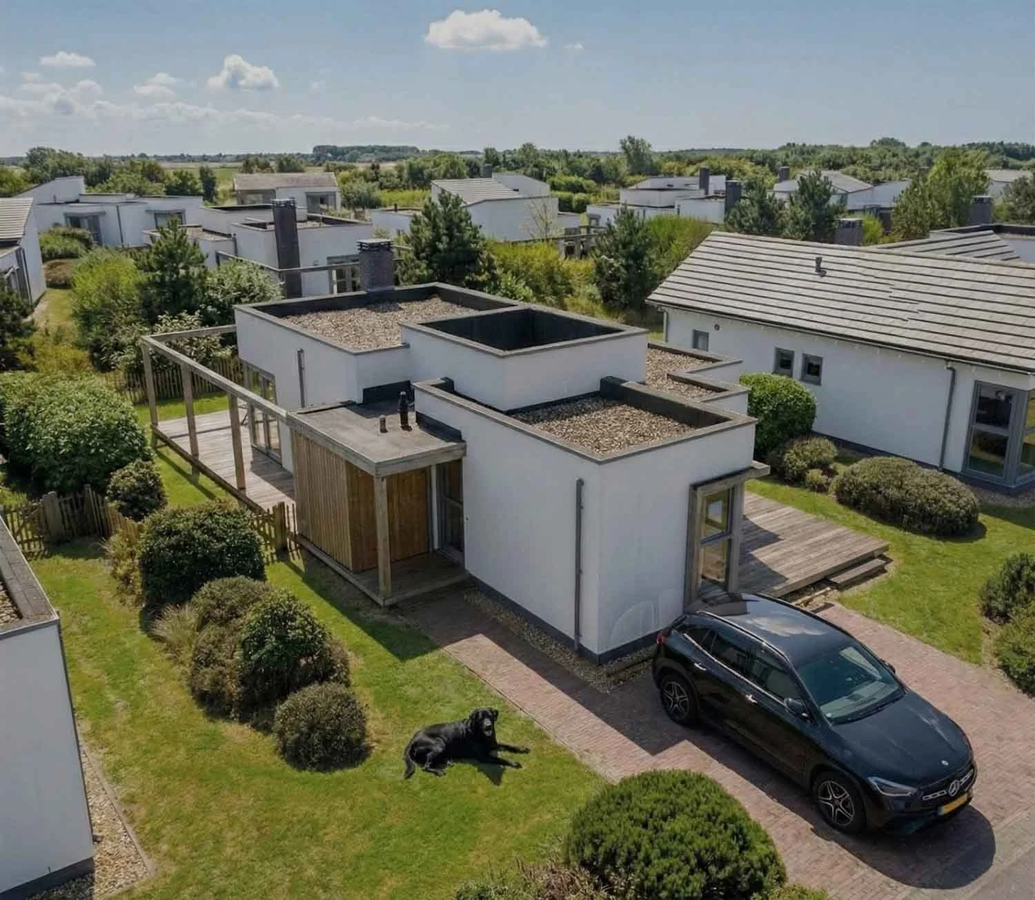 Modernes, zweistöckiges Haus mit Flachdächern, Garten und einem schwarzen Auto davor. Ein Hund liegt auf der grünen Rasenfläche im Vordergrund.