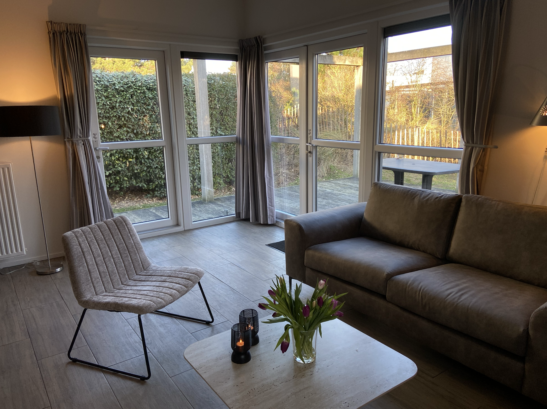 Wohnzimmer mit großer Fensterfront, Garten außerhalb, Designer-Sessel, Couch, Kaffeetisch mit Blumen und Kerzen, gemütliches Ambiente bei Sonnenuntergang