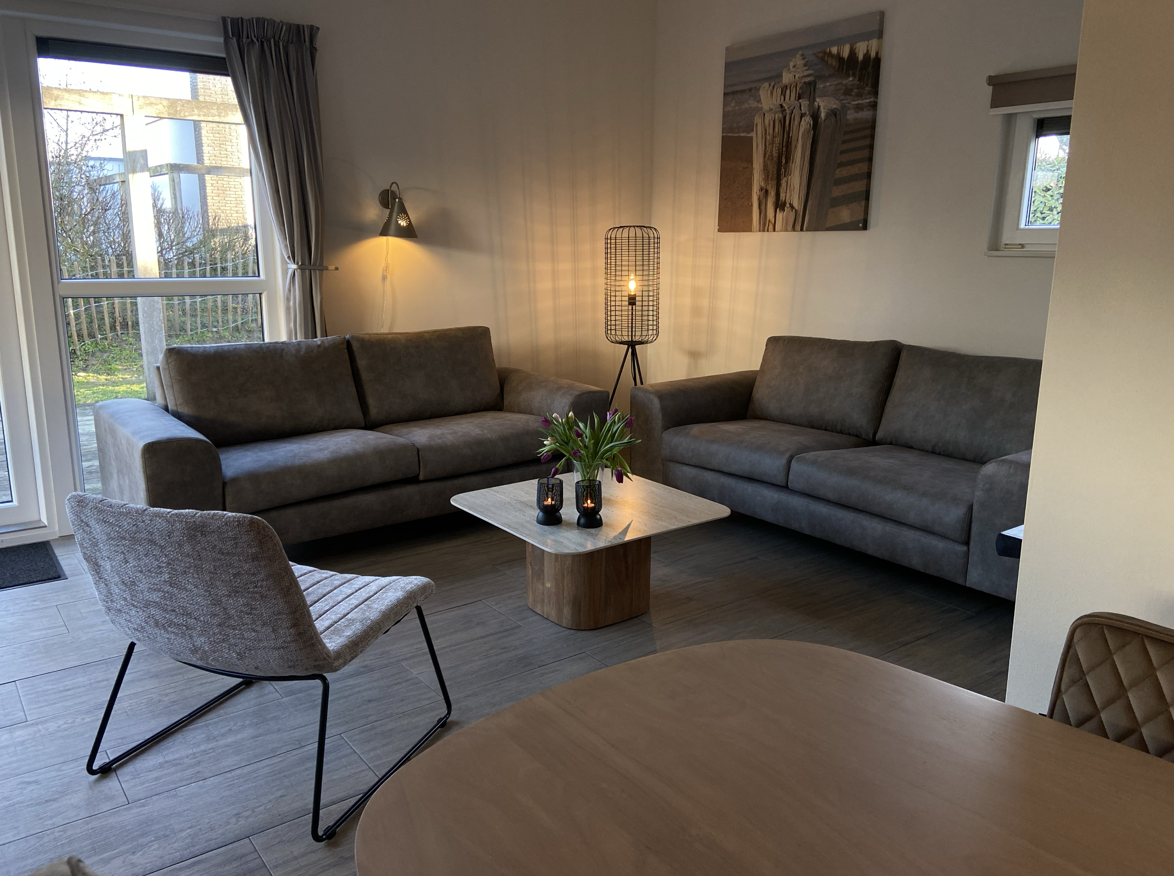 Gemütliches Wohnzimmer mit zwei grauen Sofas, einem Beistelltisch mit Blumen und Kerzen, einem Sessel, einer Stehlampe und einem großen Wandbild, viel Tageslicht durch Fenster.