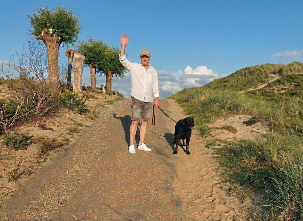 Ein Mann mit Sonnenbrille und weißem Hemd läuft einen Sandweg mit einem schwarzen Hund an der Leine entlang, im Hintergrund grüne Dünen und ein blauer Himmel.