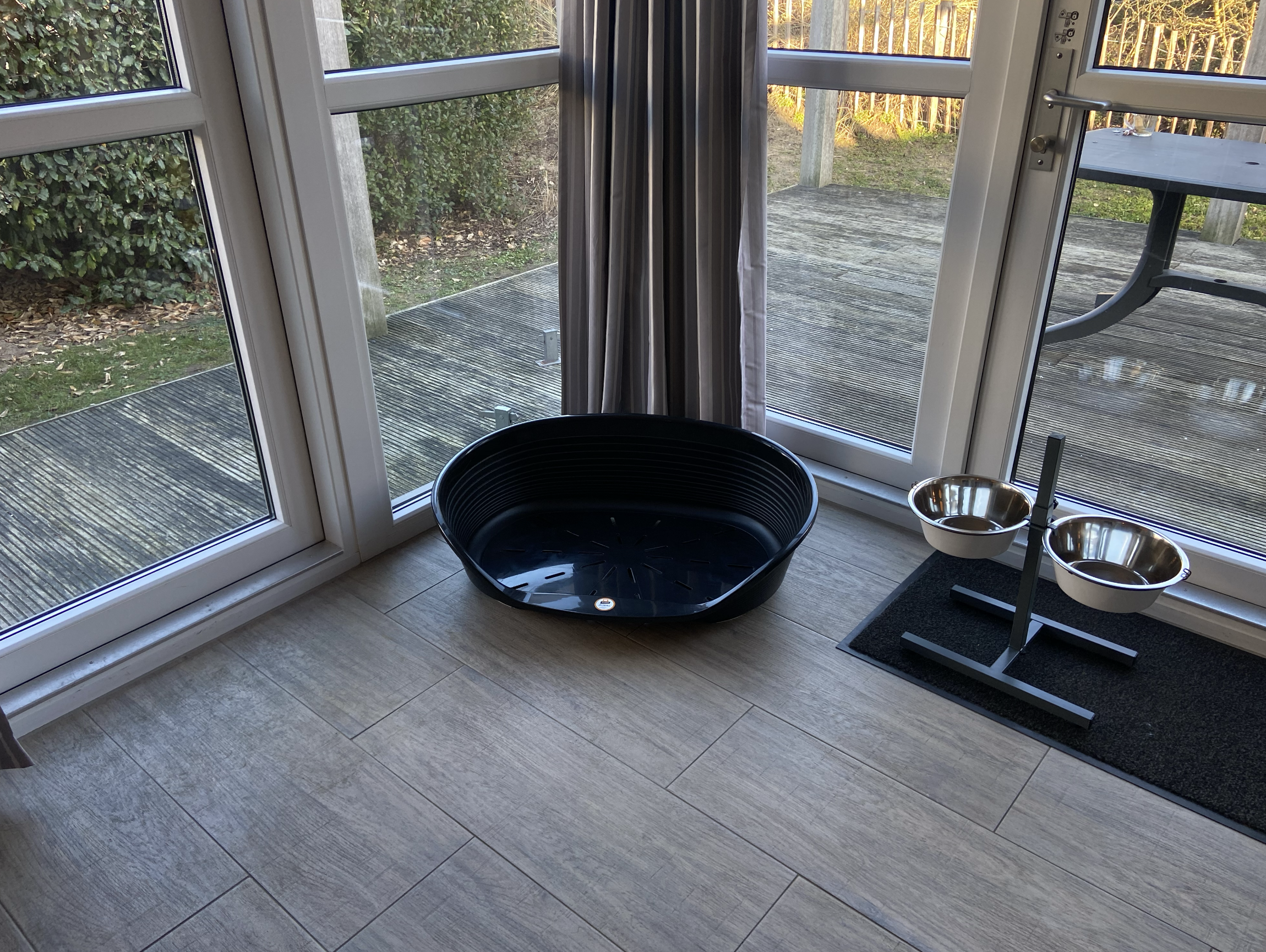 Ein leerer schwarzer Katzenkorb neben zwei metallenen Futternäpfen auf einem schwarzen Futternapfständer vor einer Glas-Schiebetür nach draußen.