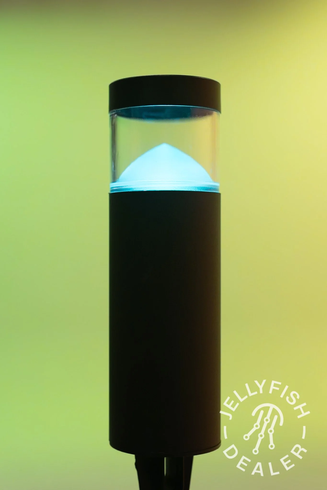 A cylindrical Jellyfish landscape Light with a black top and bottom, and a transparent middle section showing a white cone-shaped object inside, set against a yellowish-green background.