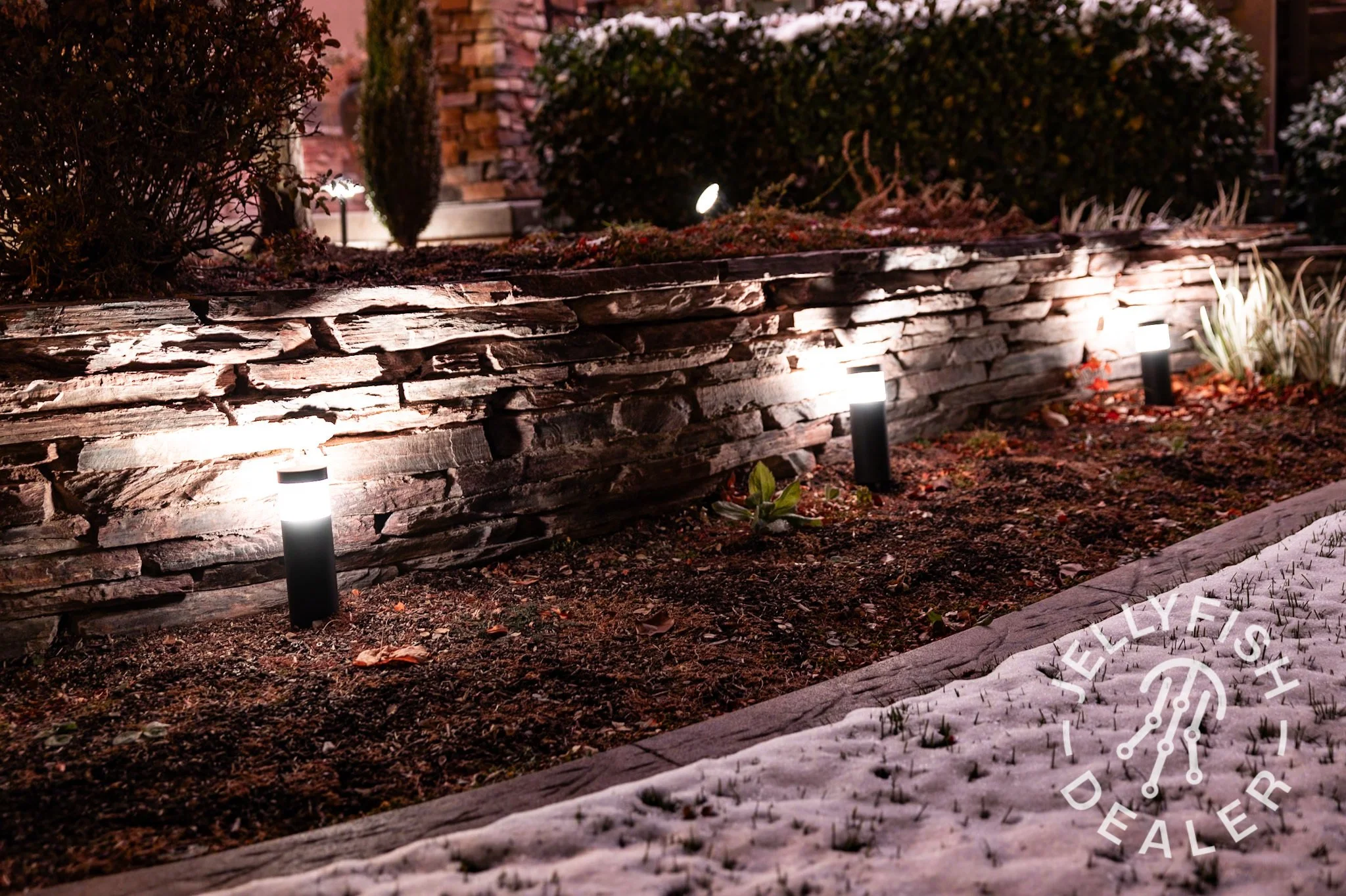 Nighttime outdoor garden scene with a stone retaining wall, garden beds with plants, and landscape lighting from Jellyfish Lighting