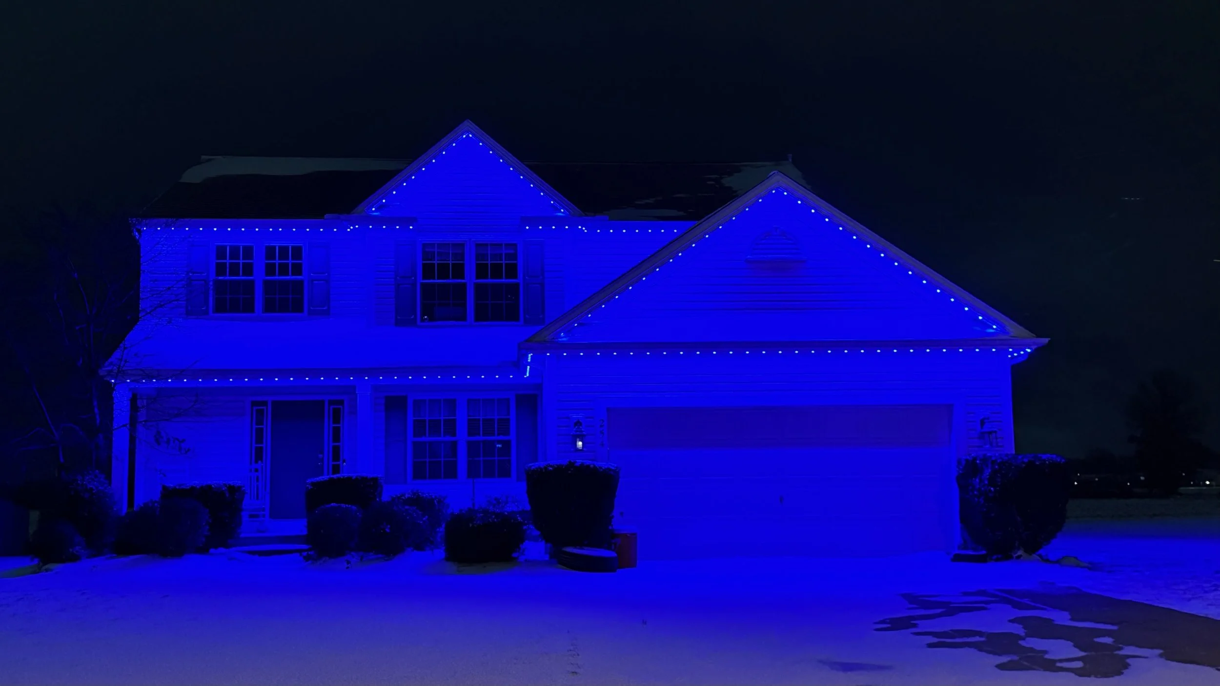 A two-story house decorated with blue Christmas lights outlining the roof, windows, and garage door at night.