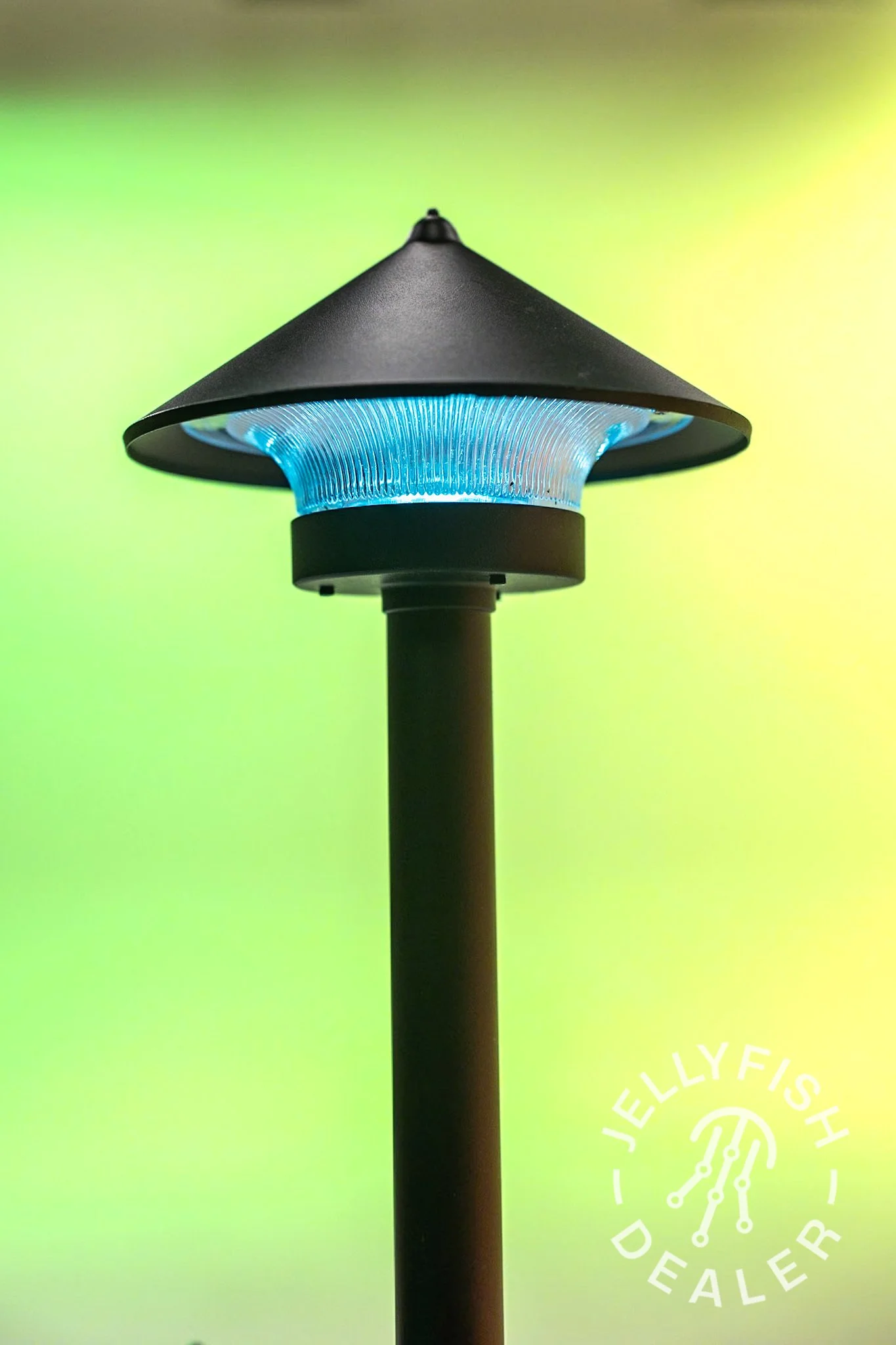 Close-up of a modern outdoor Jellyfish Landscape light with a black conical shade and a blue illuminated base, against a blurred green and yellow background.