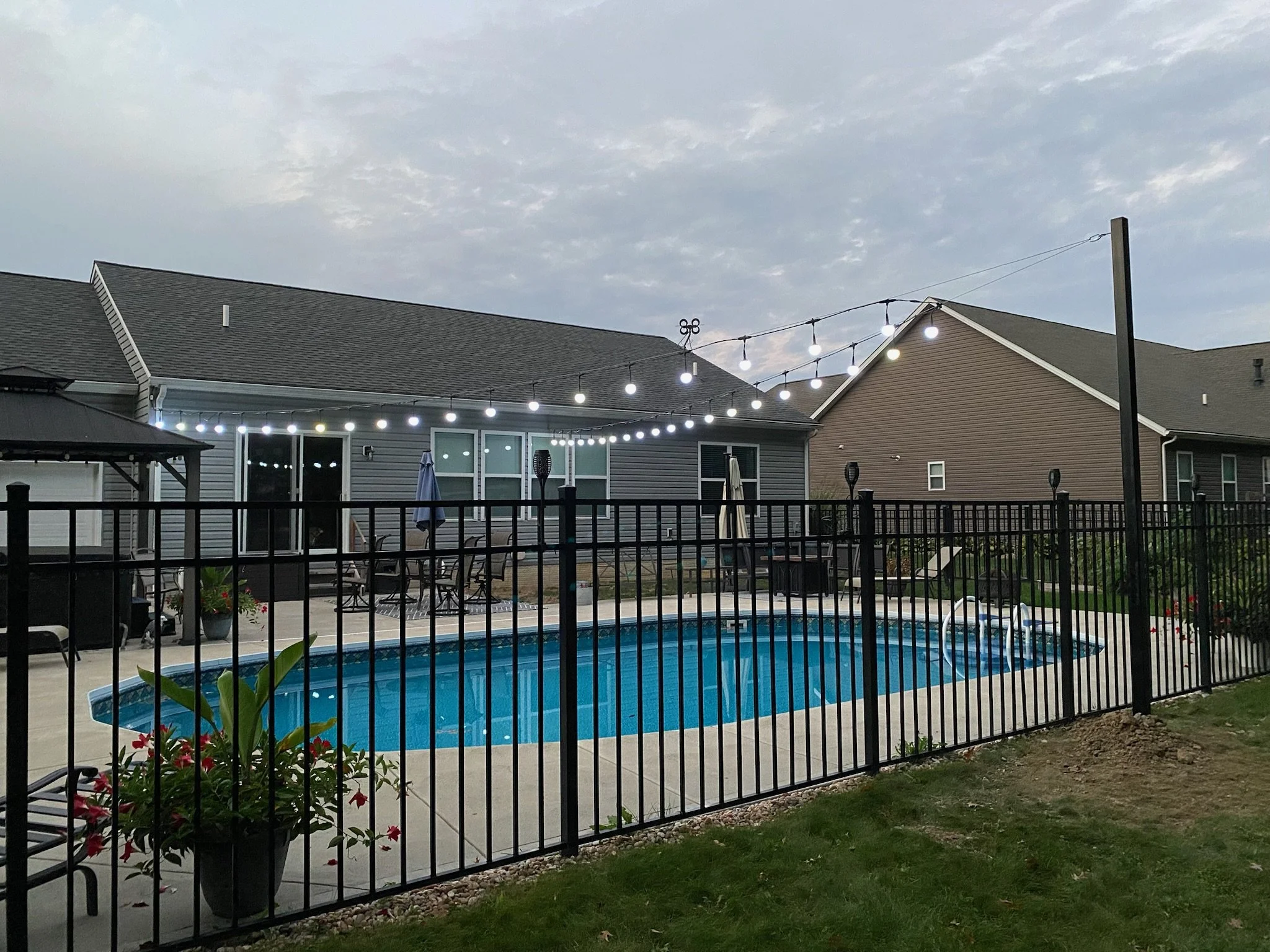 Backyard with swimming pool, patio furniture, string lights, and residential houses under cloudy sky.