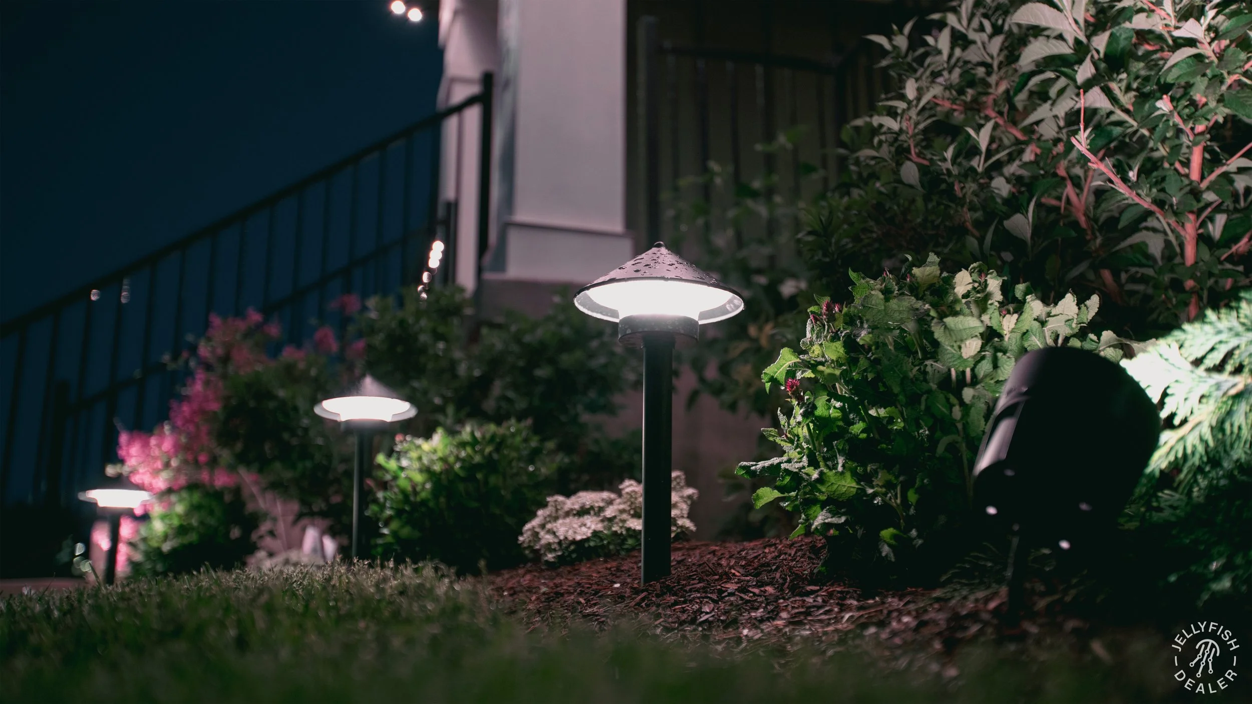 Nighttime outdoor scene with illuminated garden lamps, flowering plants, and bushes, with a structure featuring a staircase and railing visible in the background.