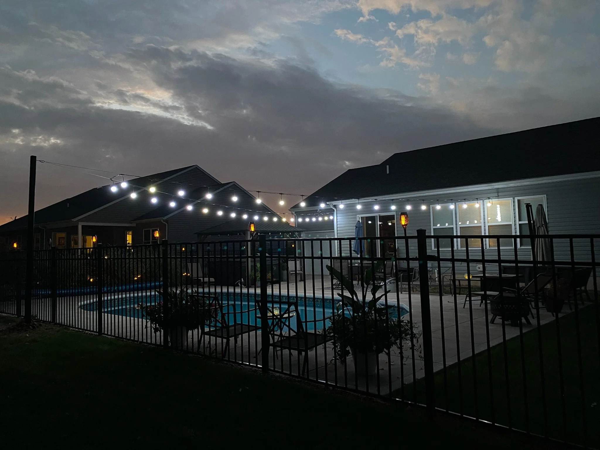 A backyard scene at dusk with a fenced swimming pool, outdoor furniture, string lights, and a house with illuminated windows under a cloudy sky.