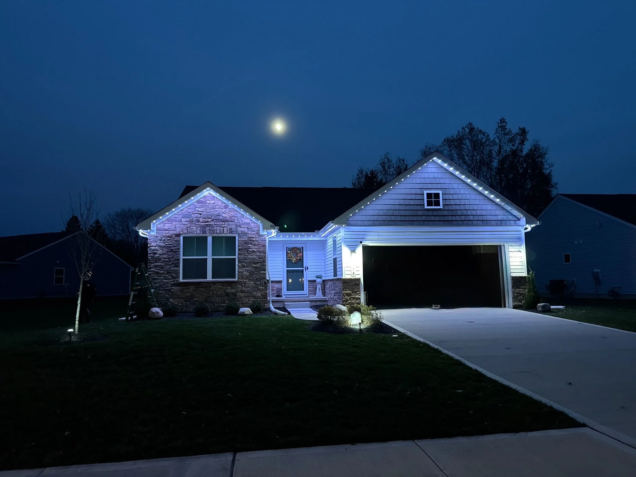 Dark night sky with a bright full moon above a suburban house decorated with string lights around the roofline. The house has a stone and siding exterior, a front porch with a door and windows, and a driveway with some bushes and trees in the yard.
