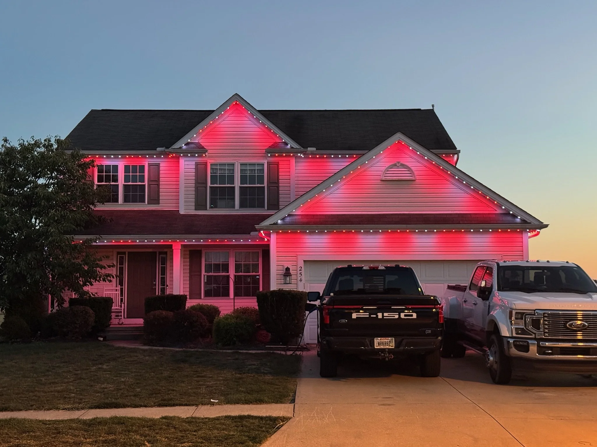 A two-story house decorated with pink and red Christmas lights on the roof and around the windows. Two trucks are parked in the driveway, and the sky is transitioning from day to night.