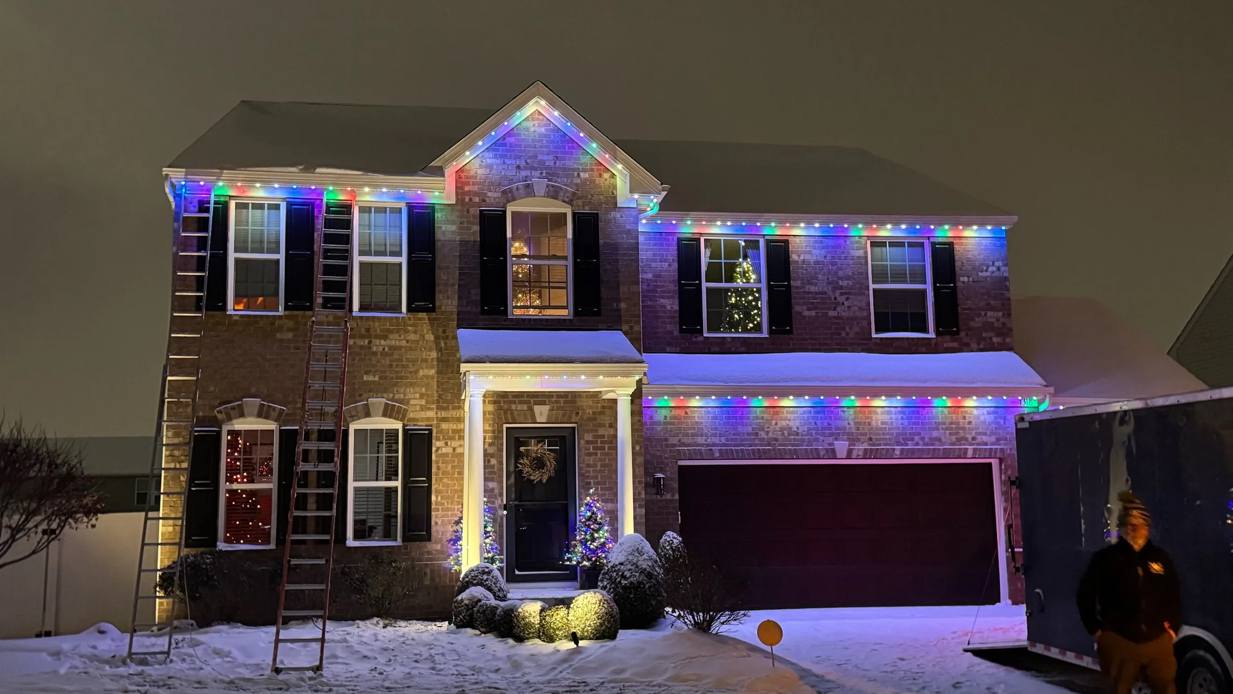 A two-story brick house decorated with colorful Christmas string lights along the roofline and around windows, with a Christmas tree visible through the window, snow covering the yard, and a person standing near the truck on the right side of the ima