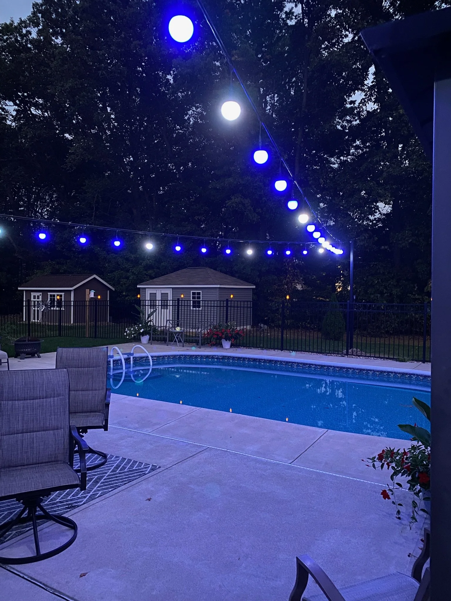 Nighttime backyard scene with a swimming pool, patio furniture including a table and chairs, potted plants, and string lights with blue and white bulbs hanging overhead, with trees in the background.