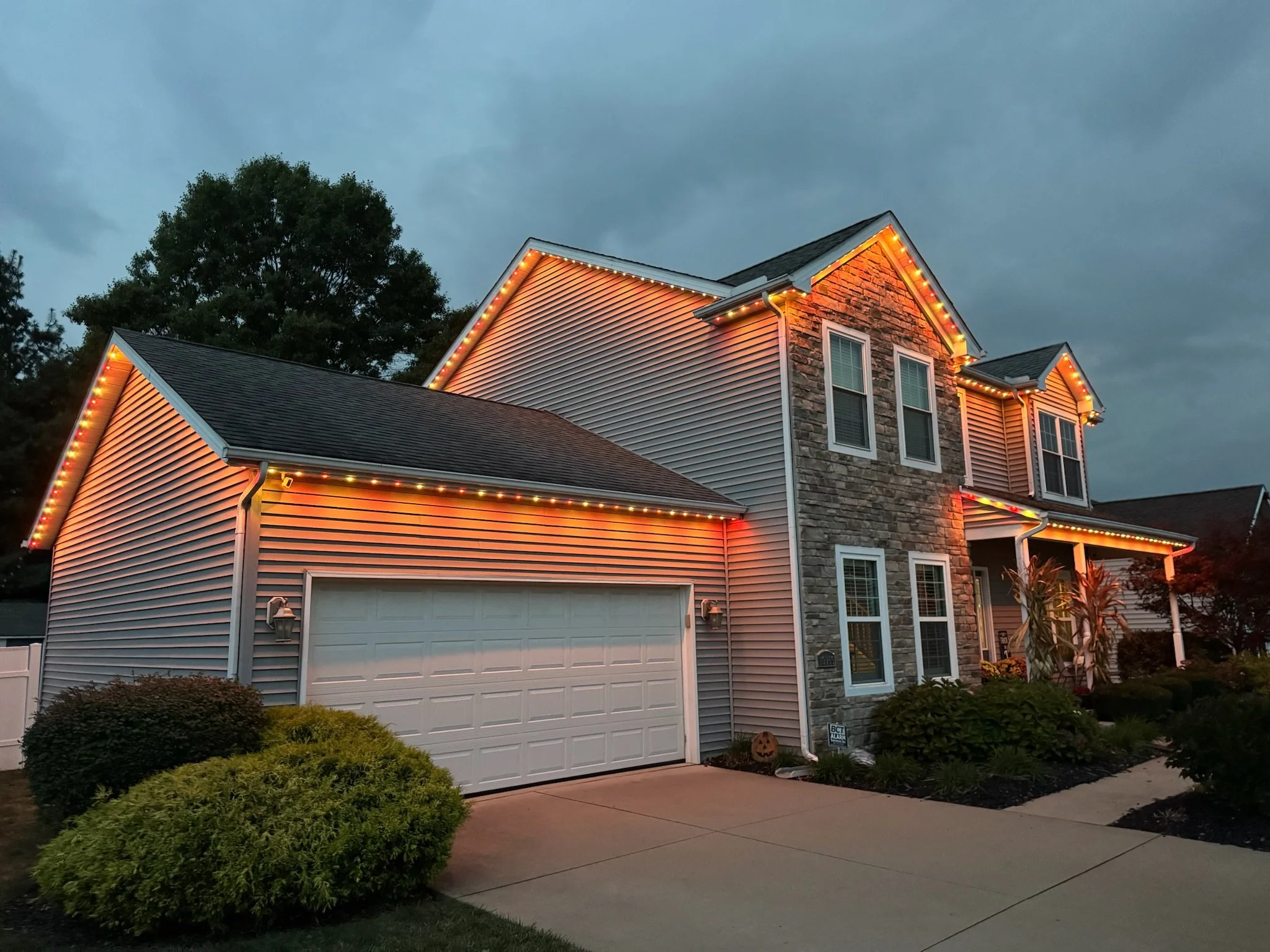 A two-story house decorated with string lights along the roofline in the evening, with a large tree and a cloudy sky in the background, a garage door, and a well-maintained garden with bushes and a pathway in the front yard.