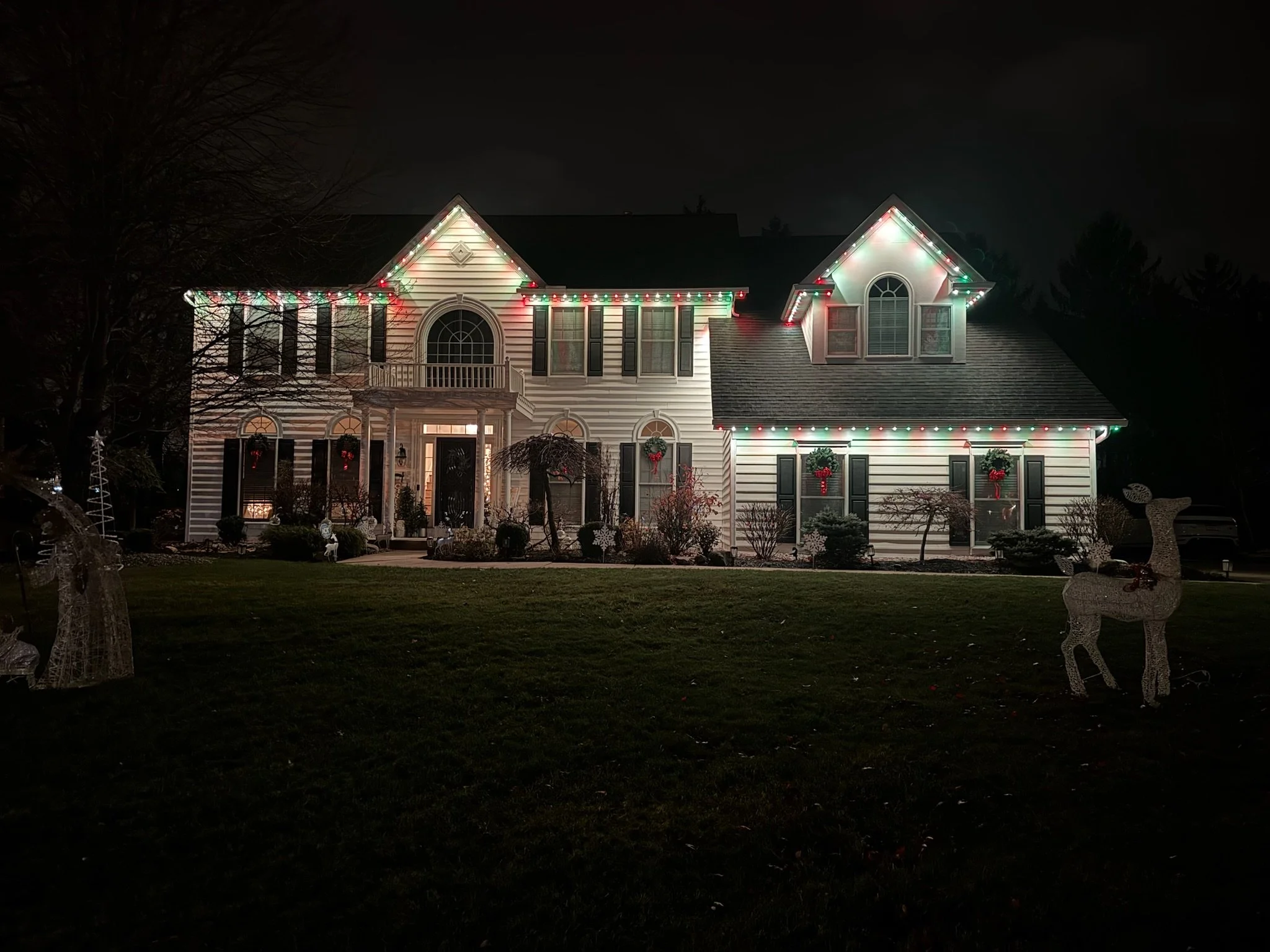 A house decorated with Christmas lights at night, with windows and porch ornaments. The decorative lights are primarily red and green, outlining the house's roof and windows.