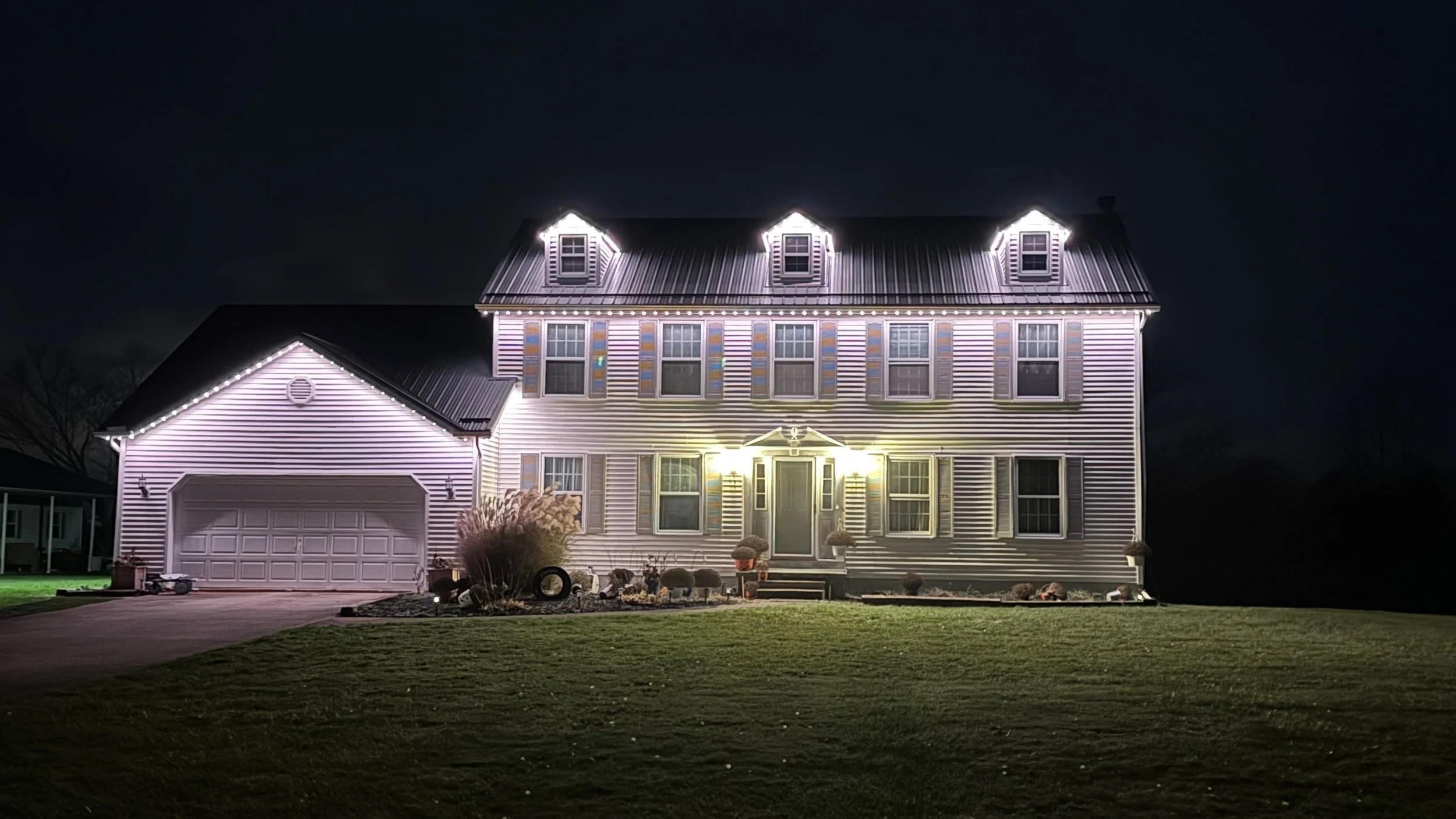 A large, well-lit house at night decorated with Christmas lights along the roofline, windows, and porch.”}