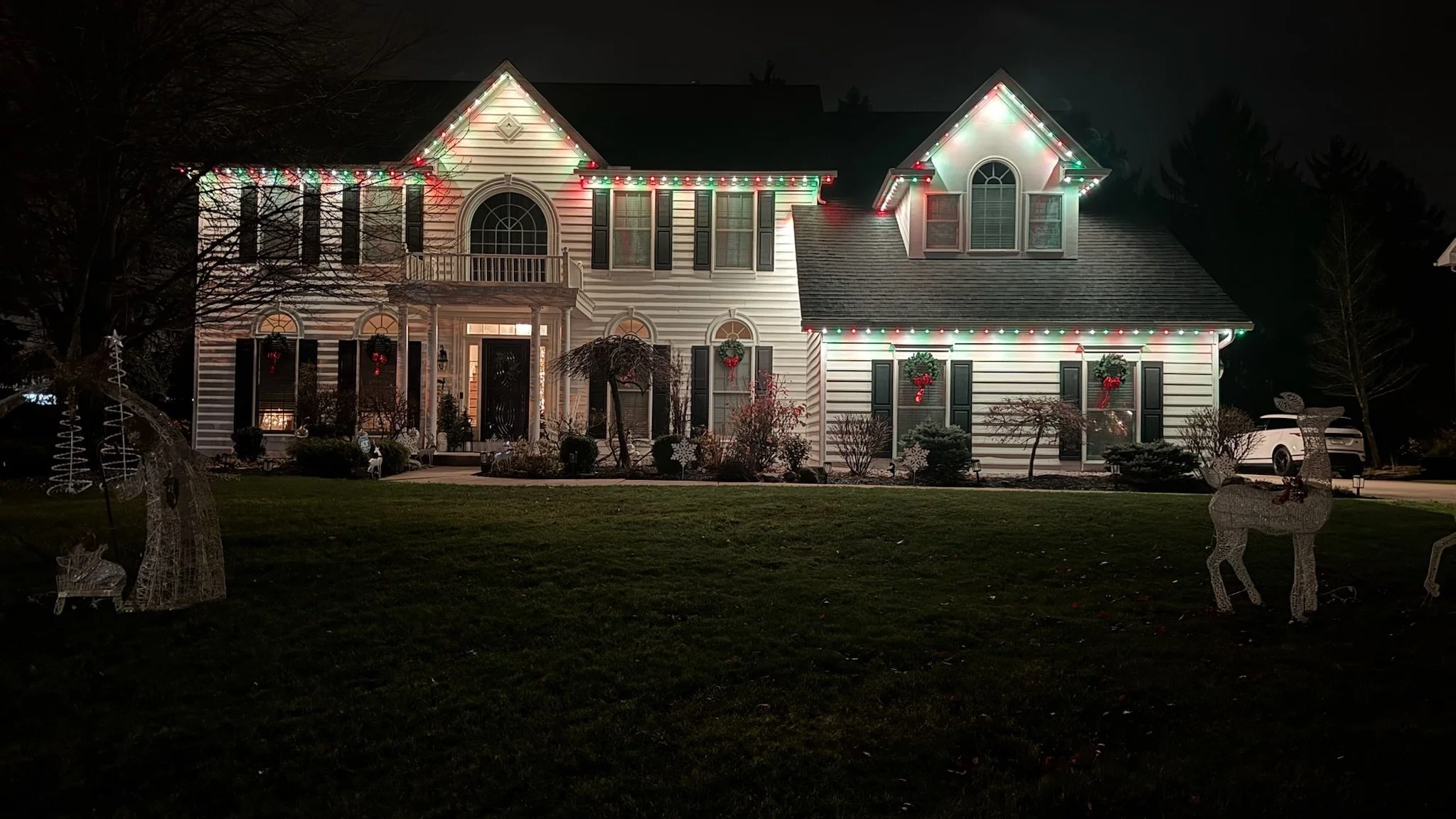 Night view of a house decorated with Christmas lights and holiday decorations, including reindeer figures and wreaths on windows.