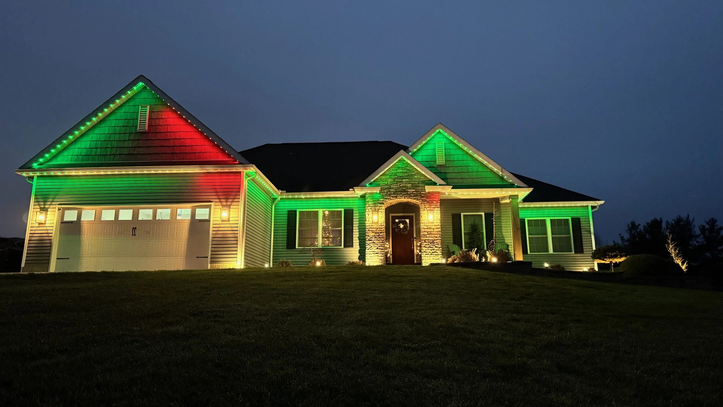 A house decorated with colorful holiday lights at night, with green, red, and white lights outlining the roof and windows.