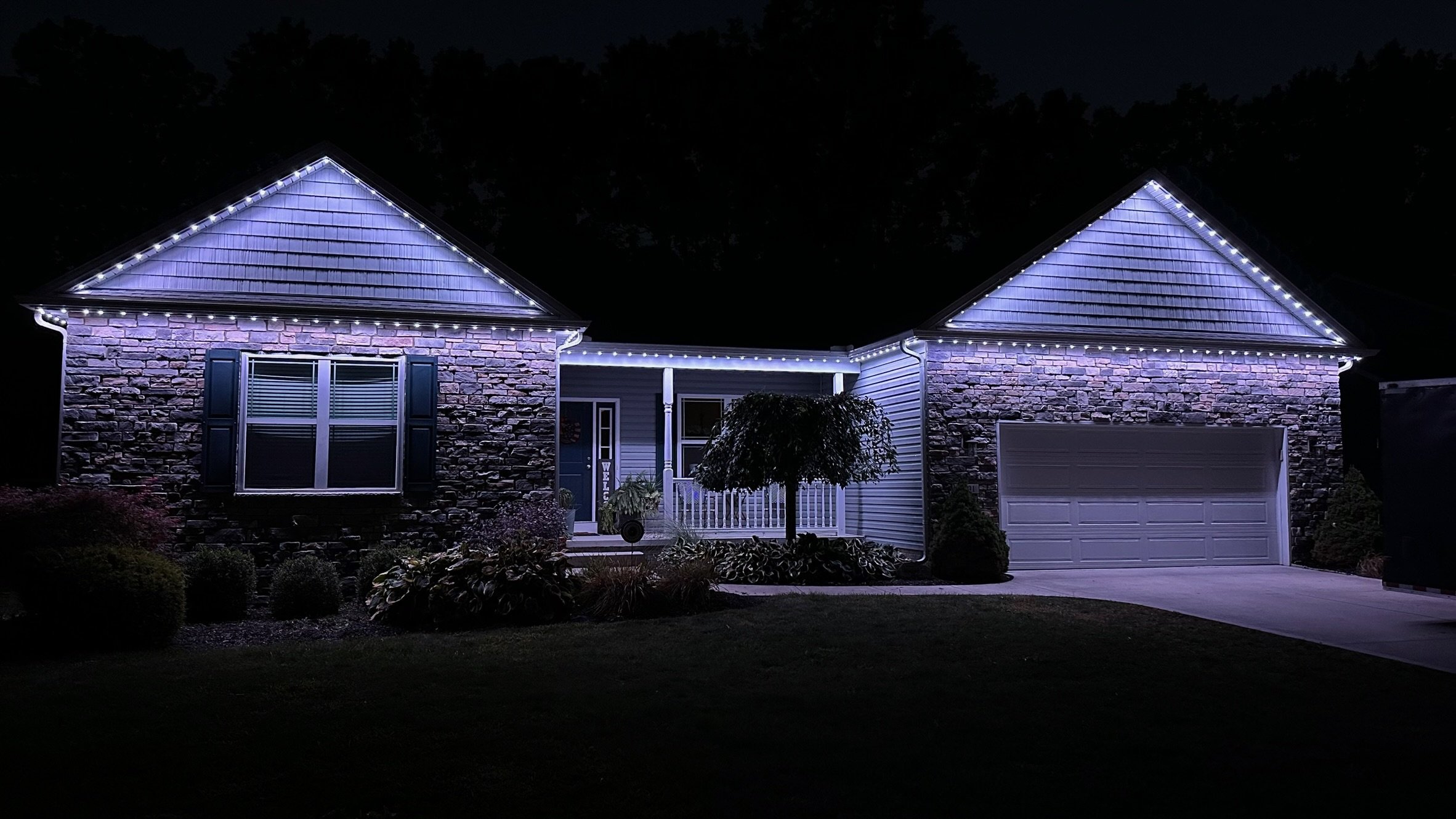 A house decorated with white Christmas lights outlining the roof and eaves, with a stone and siding facade, a front window with shutters, a small porch, and a garage door. The yard has bushes and a small tree, illuminated by the house lights at night