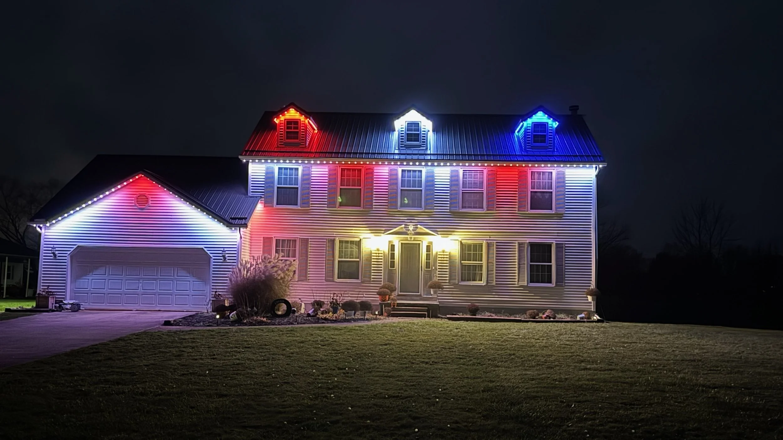 A house decorated with colorful Christmas lights at night, featuring red, white, and blue lights on the roof and around the windows.