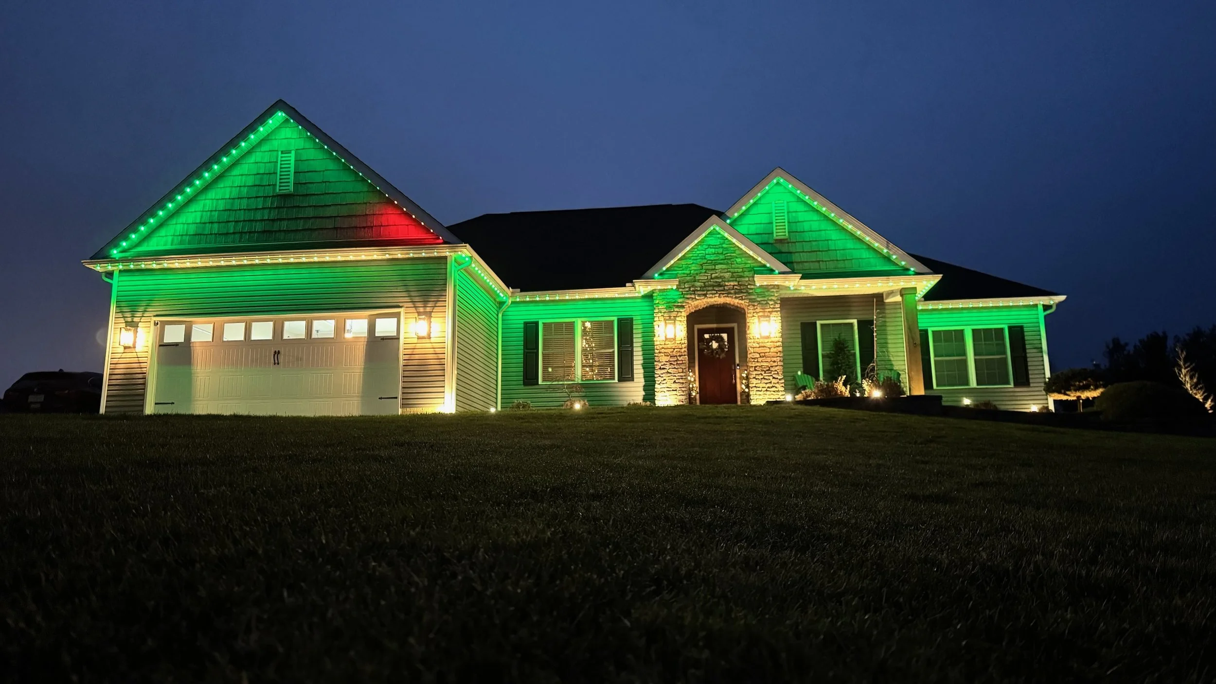 A house decorated with green and red Christmas lights at night, with a dark sky in the background and a well-maintained lawn in the foreground.