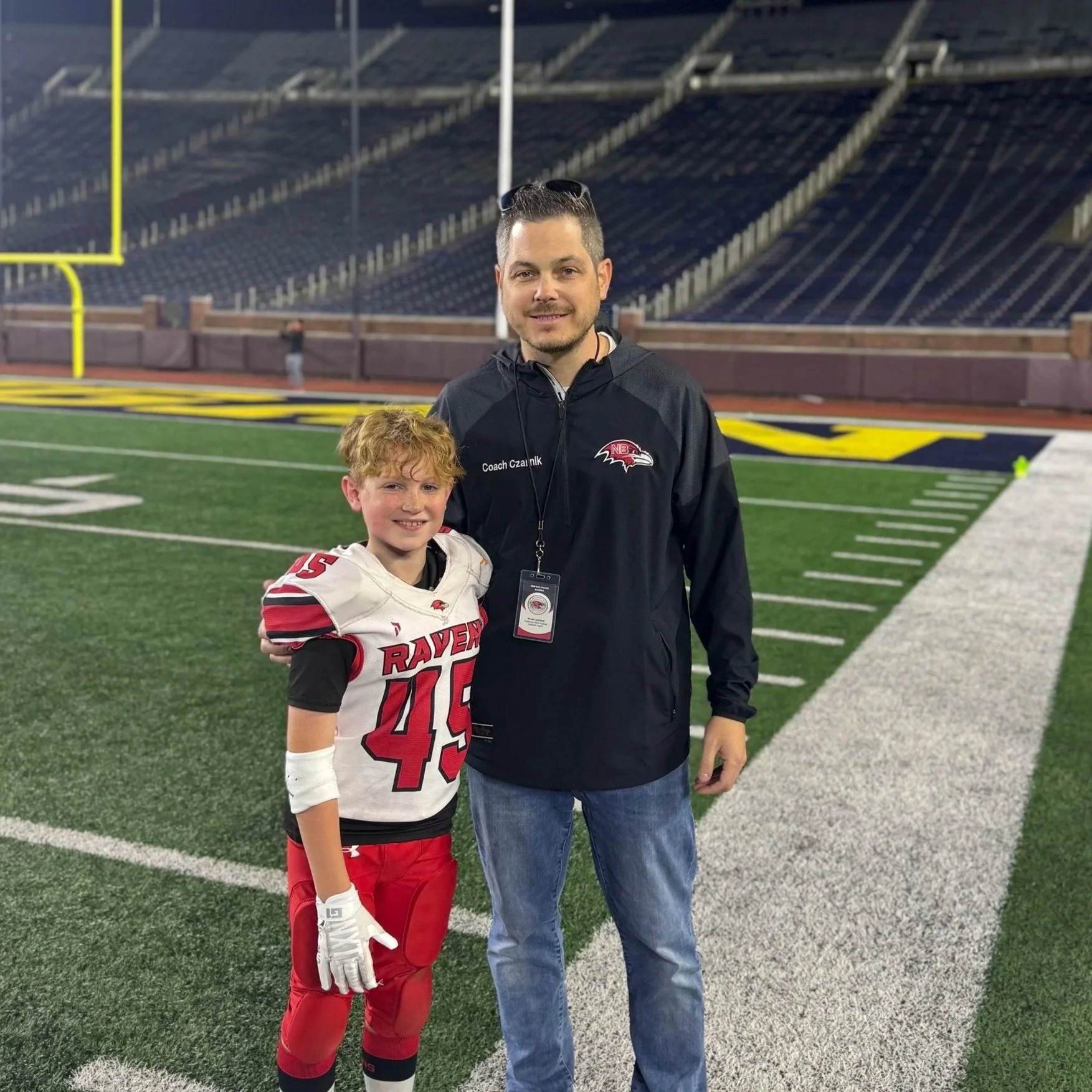 A coach and a young football player standing on a football field at a stadium, smiling at the camera.