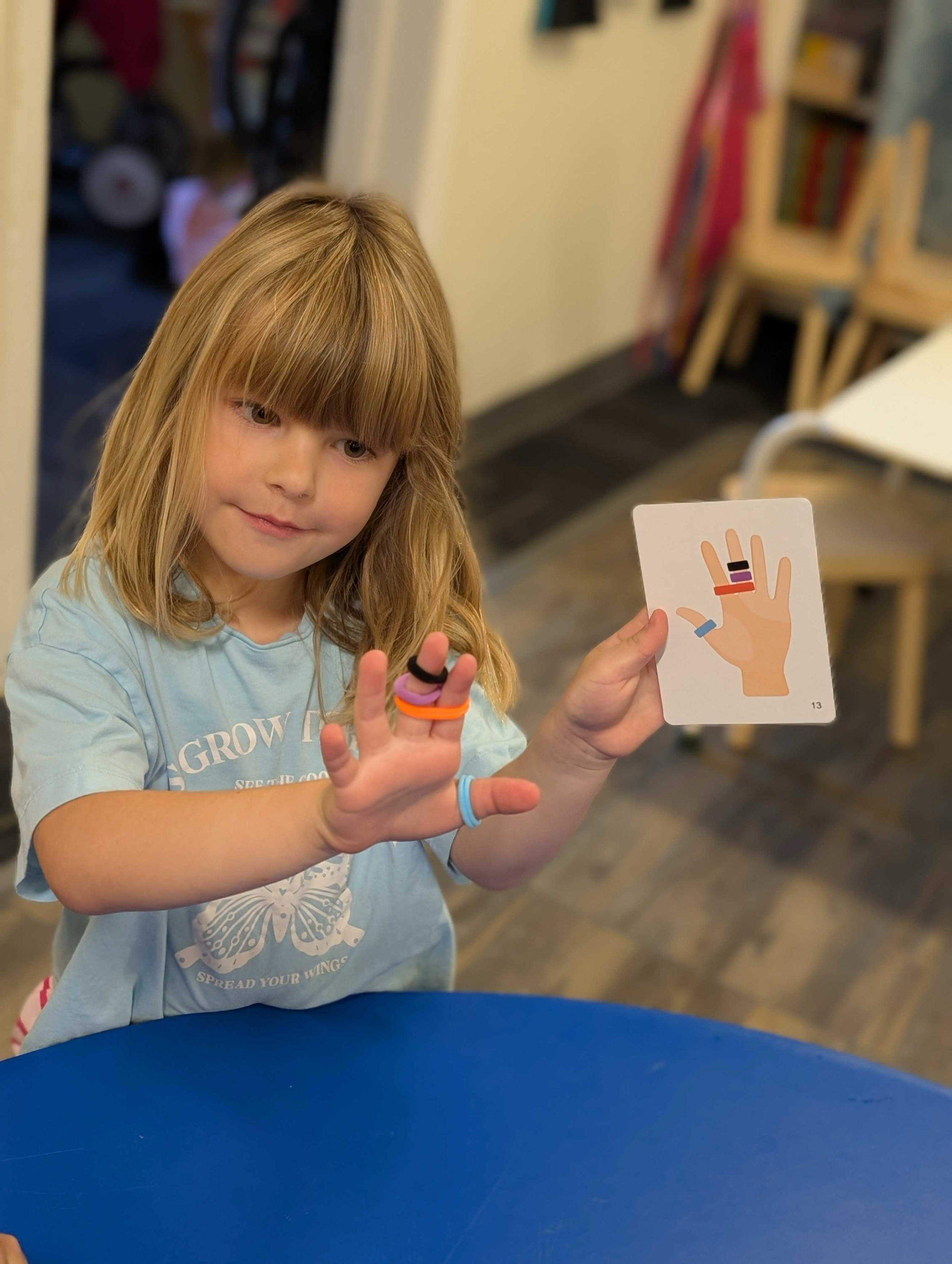 A young girl with blonde hair matching rubber bands on her fingers to those on a picture card she is holding.
