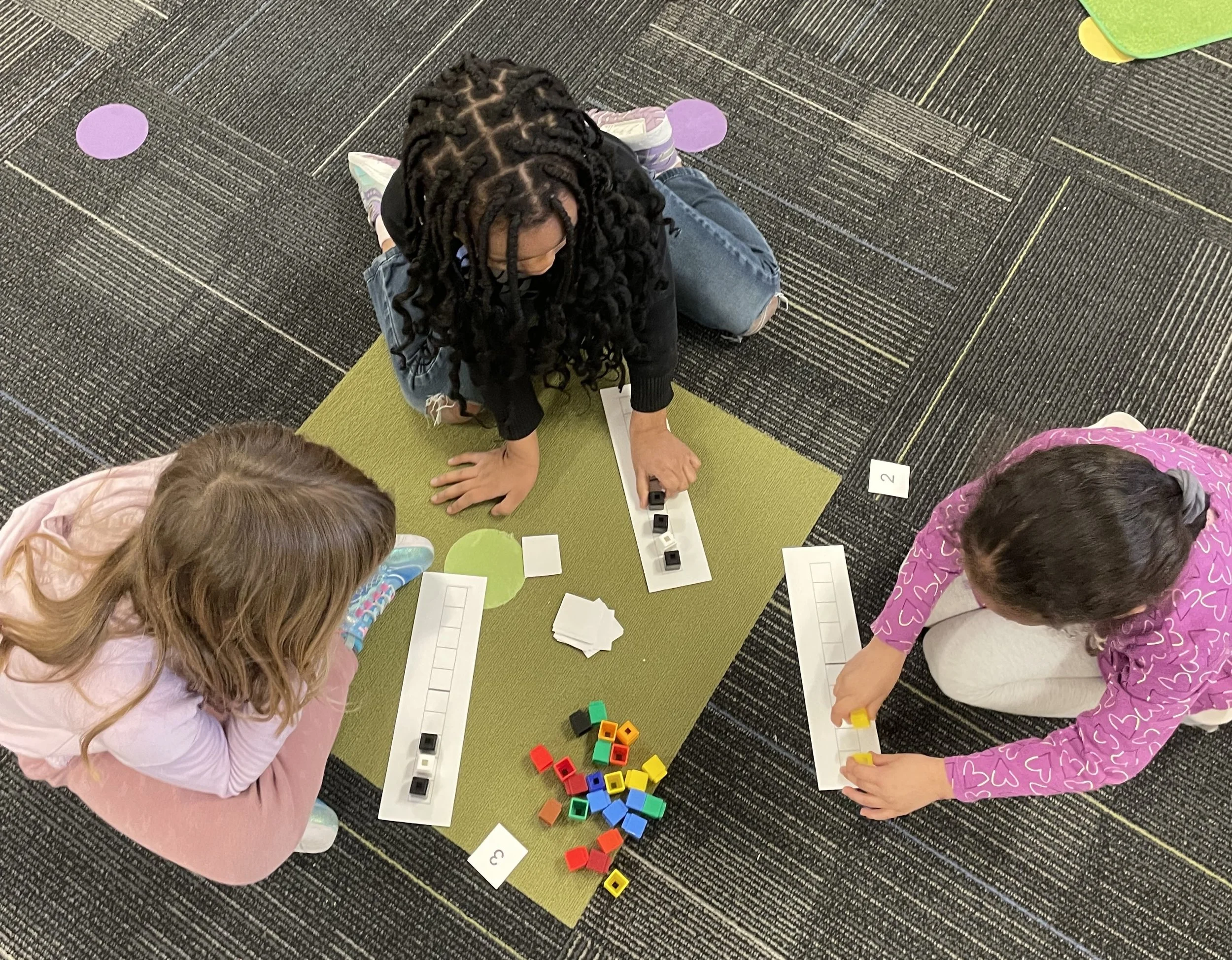 Three children playing an educational game with small cubes and numbered chart on a green mat on the carpeted floor.