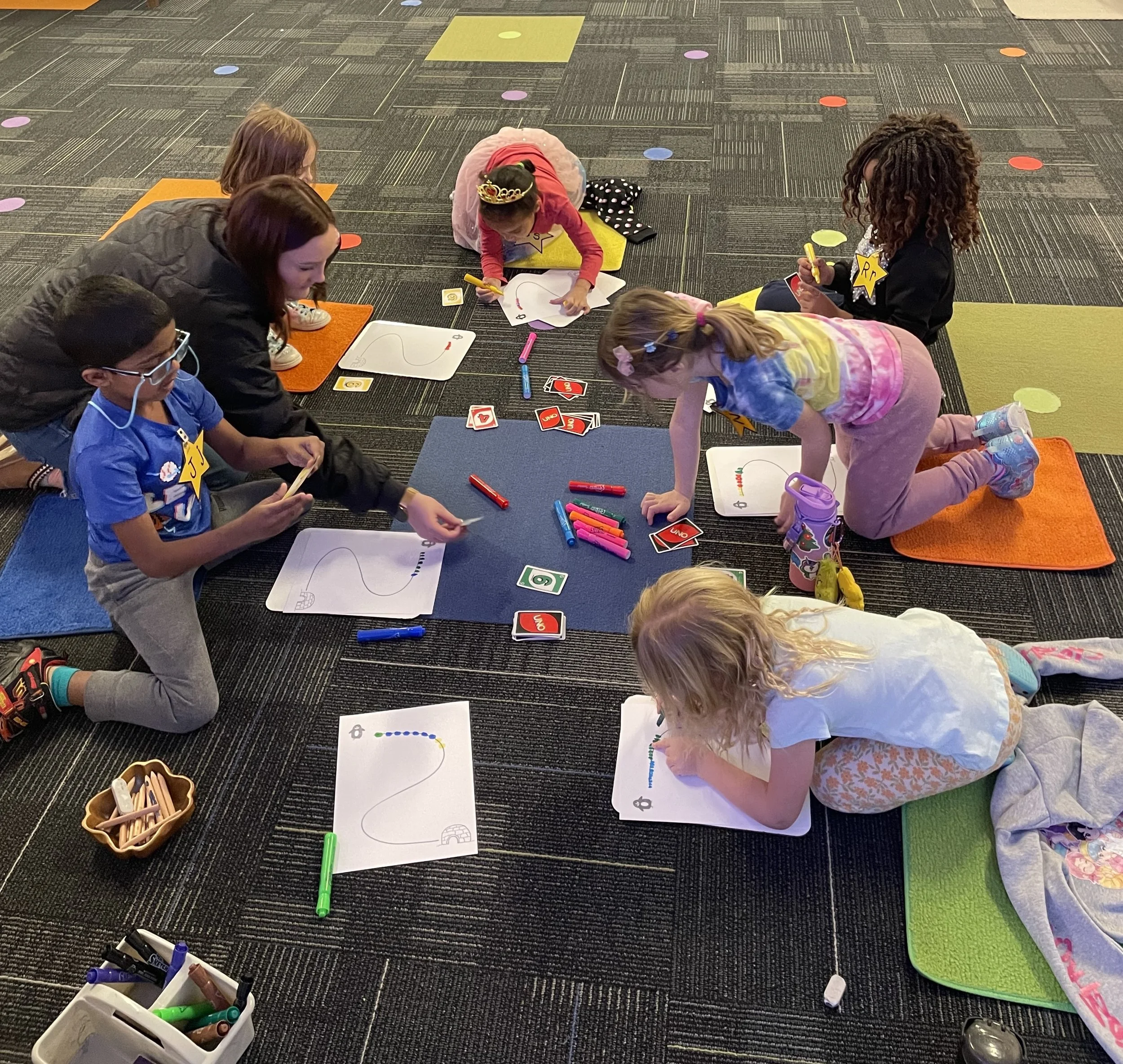 Children participating in an arts and crafts activity indoors, drawing and coloring on paper with markers, while sitting or kneeling on colorful mats.
