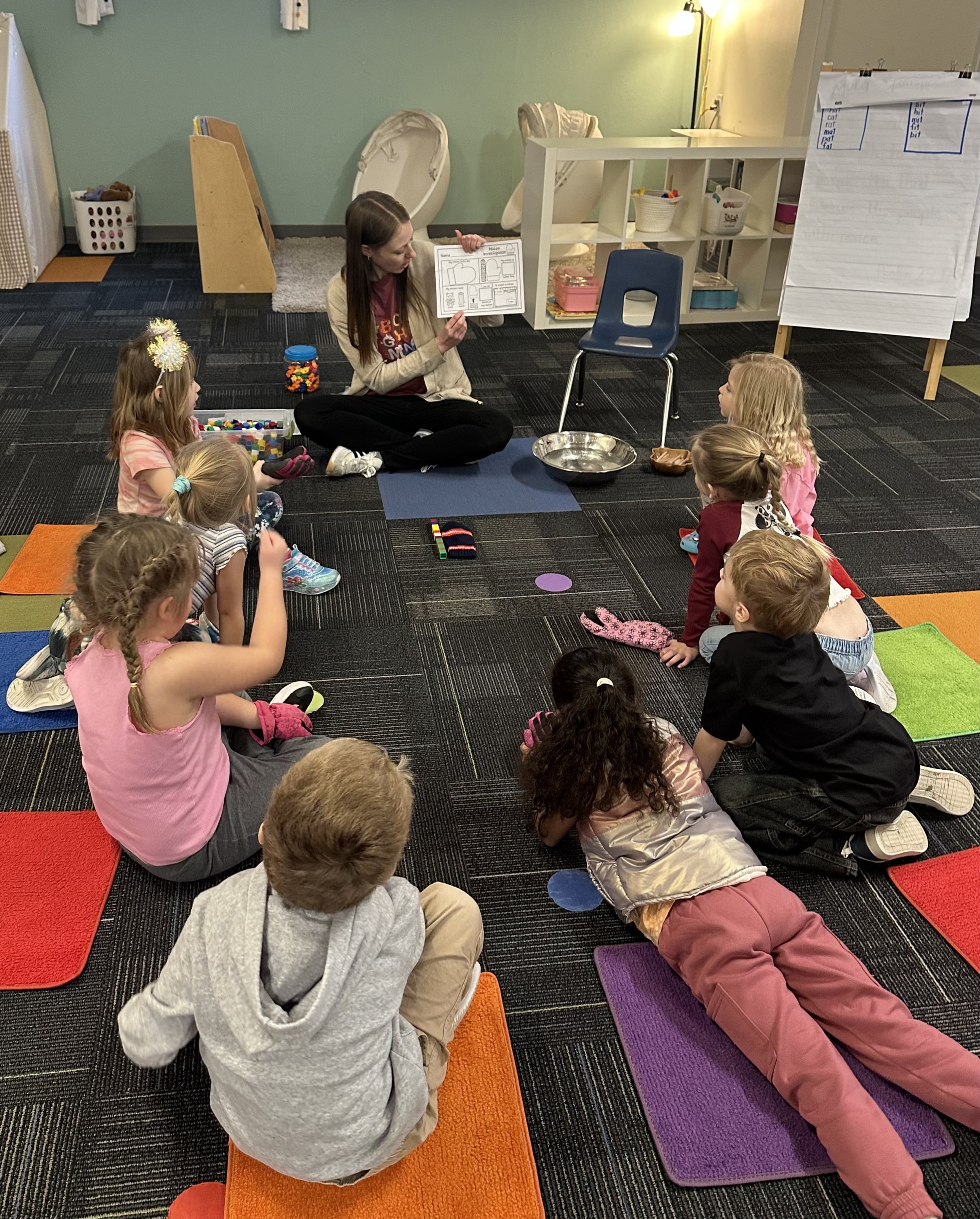 A woman teaching a group of young children in a classroom, sitting on yoga mats, with a chart, a basket of toys, and two bassinets in the background.