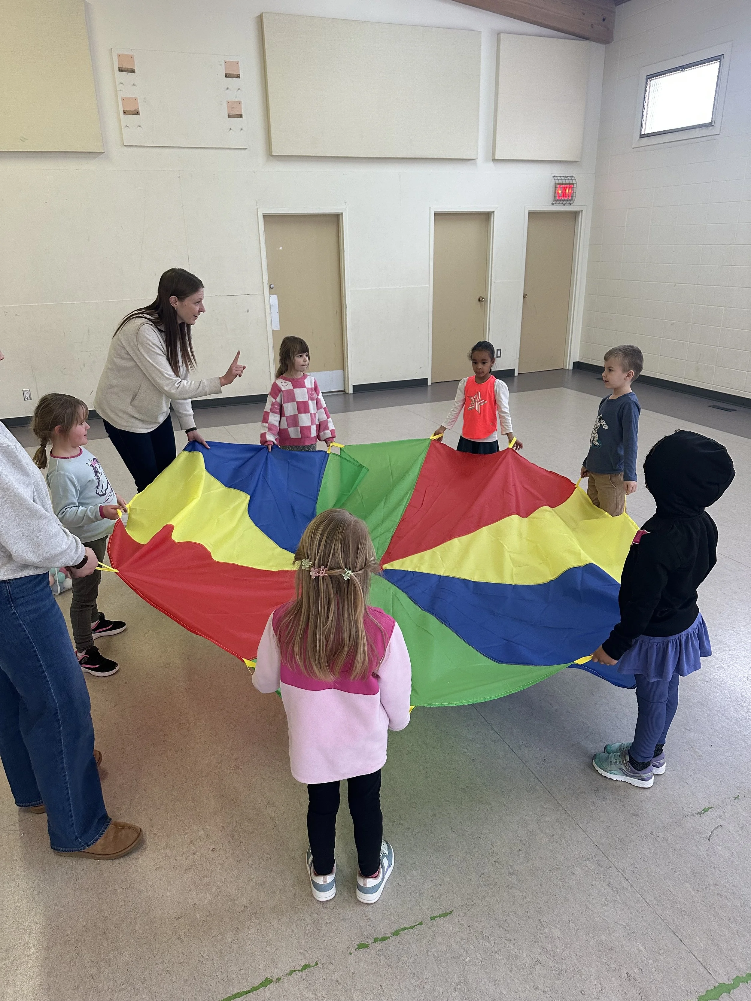 Children and adults holding a colorful parachute in a room with beige walls and tiled floor.