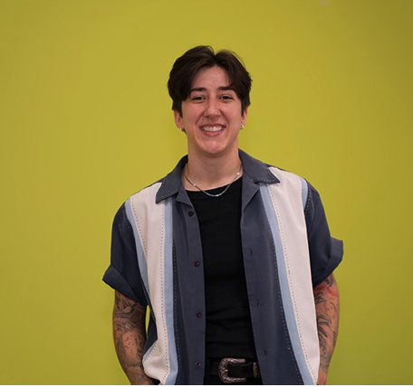 Kelsi Creek and woman with short dark hair smiling, wearing a black shirt and a denim short-sleeve button-up over it, standing against a bright yellow background for Central Health Interview